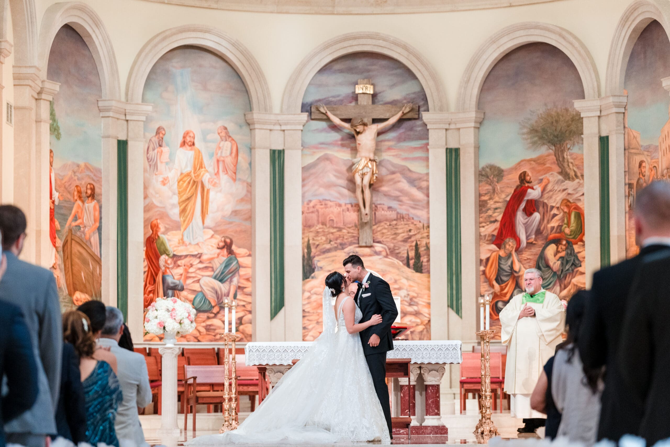 Wide shot capturing the big kiss at the altar of St. James Cathedral, with the priest, guests, and cathedral artwork in the background.
