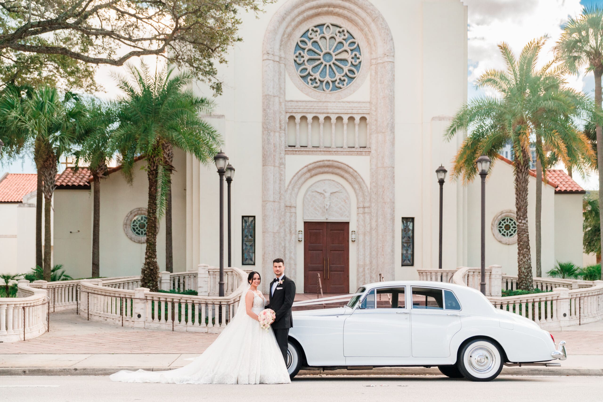 Wide shot capturing Jessica and Javier smiling at the camera in front of their car, with St. James Cathedral and an antique car in the background.