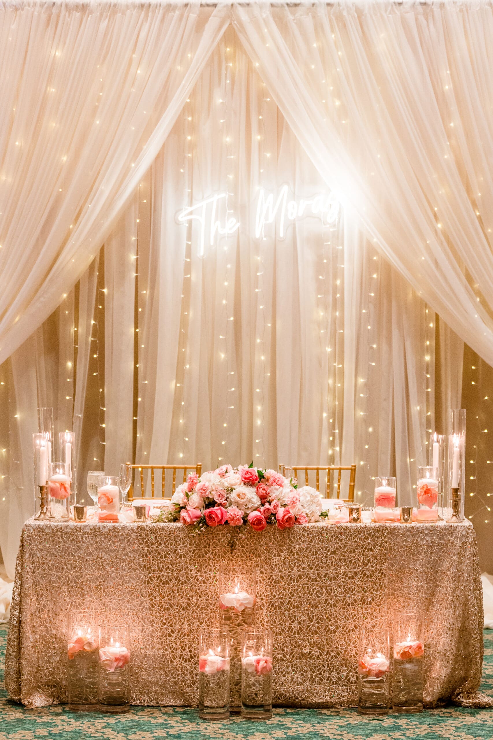 The couple's table adorned with flowers, lights, and white drapes in the background, featuring a gold-patterned tablecloth and elegant decor.