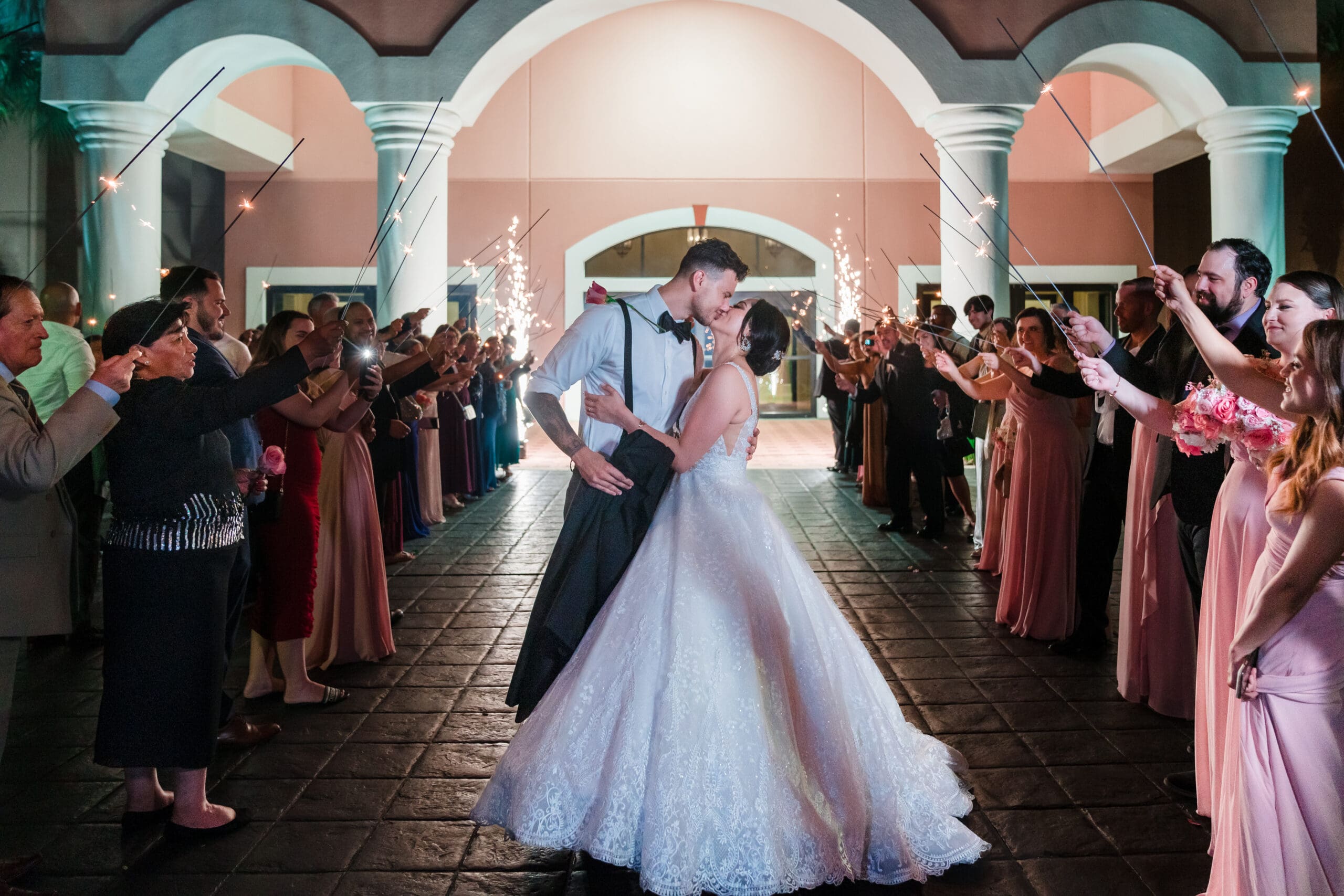 Wide shot capturing Jessica and Javier's kiss at the end of the grand exit, with guests raising their arms and holding extra-large sparklers, creating a luminous display of celebration.