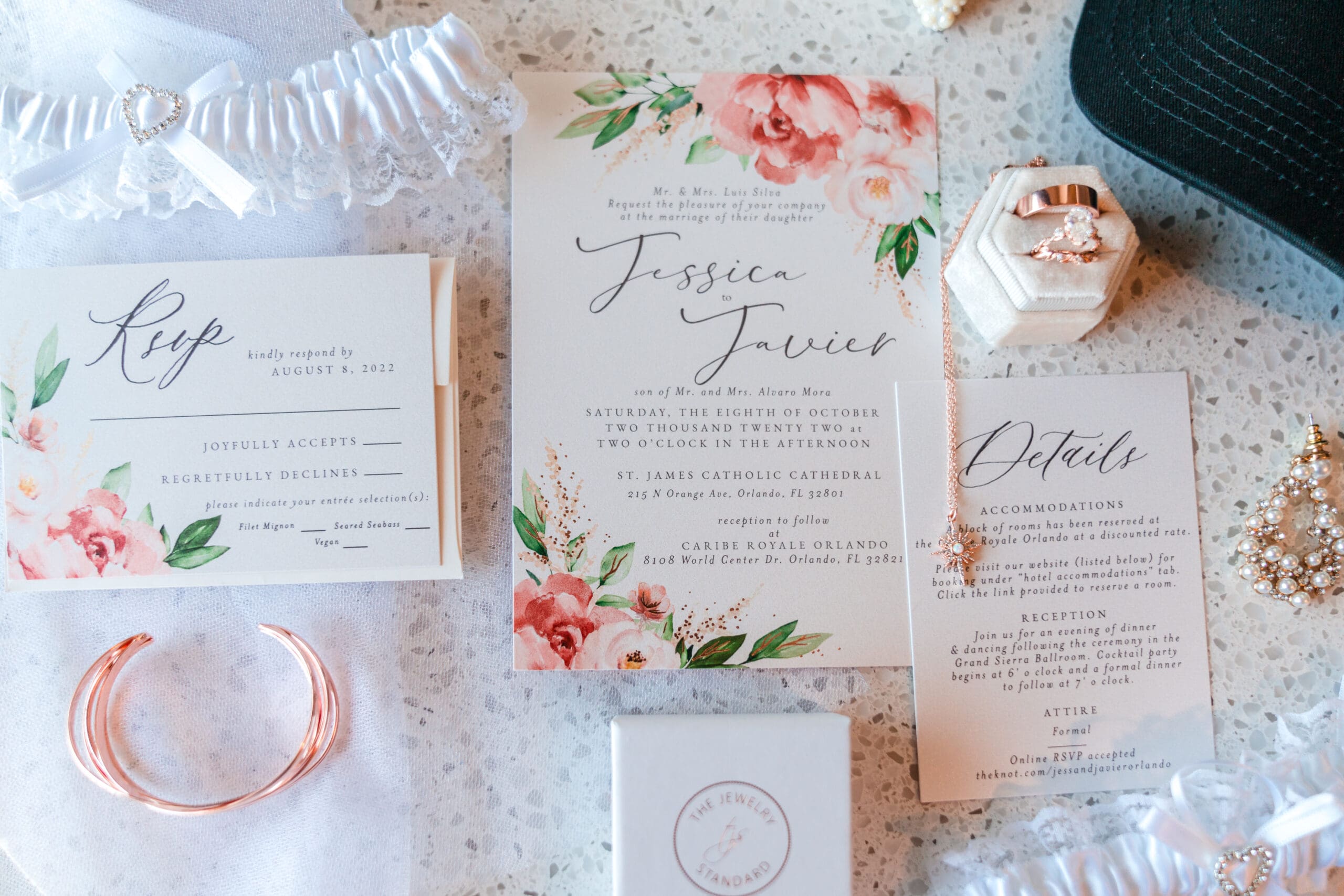 Close-up shot of wedding rings, bands, and stationary including RSVP details arranged on a table.
