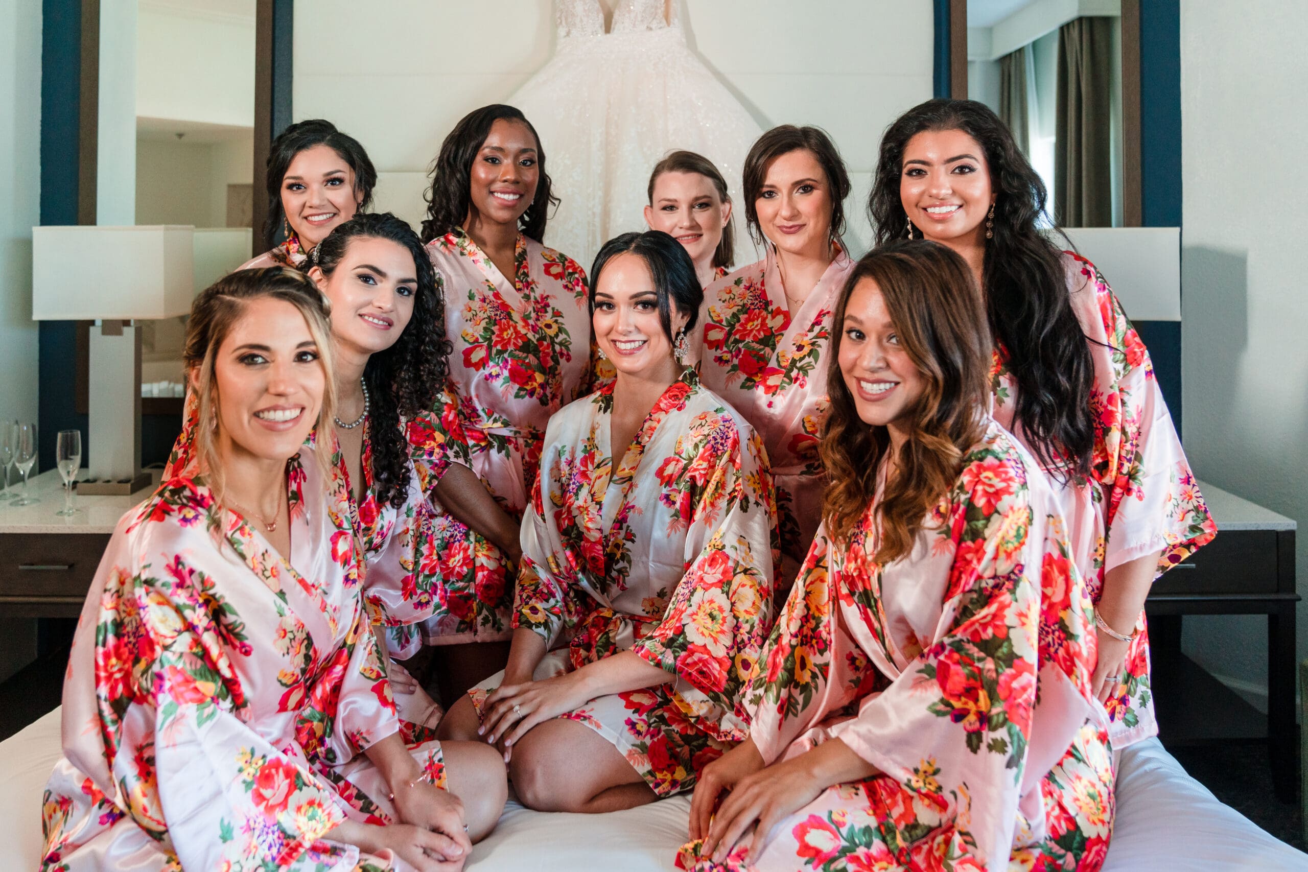 Jessica and her bridesmaids in matching flower print robes, sitting on the bed at Caribe Royale Orlando, with Jessica's wedding dress hanging in the background.