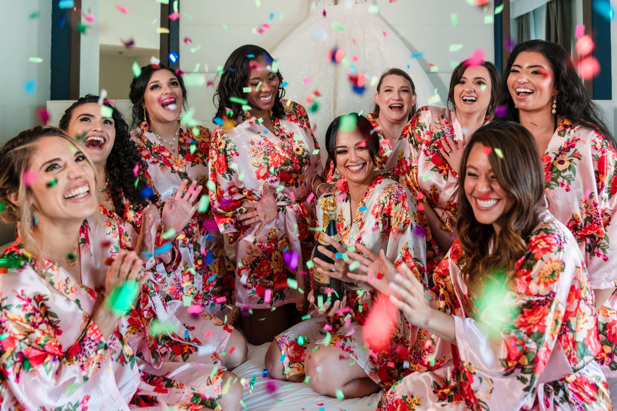 Jessica and her bridesmaids in matching flower print robes, sitting on the bed at Caribe Royale Orlando, with Jessica's wedding dress hanging in the background. Confetti and champagne bottles are seen around them.