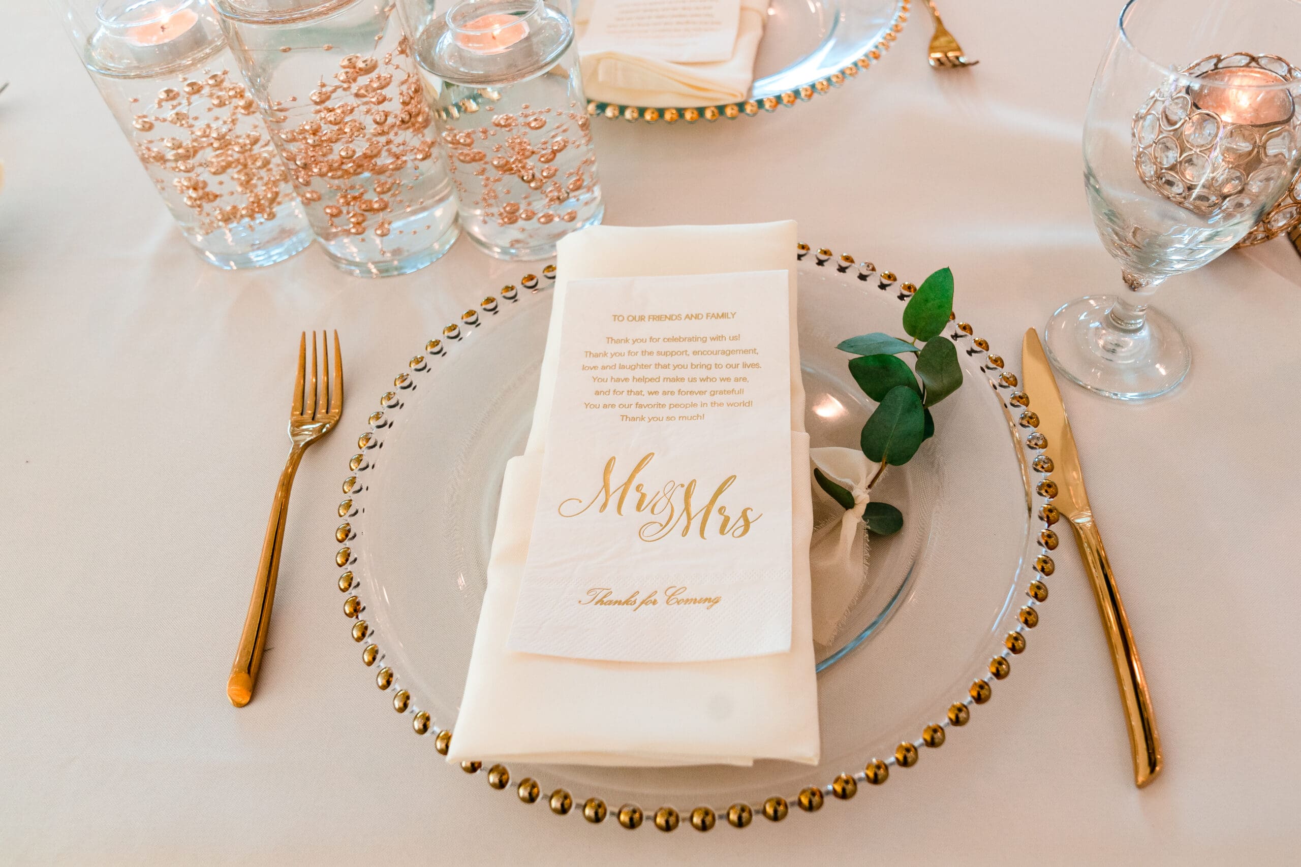 Close-up of elegant table arrangement with gold silverware, gold-rimmed white plates, and napkins with 'Mr. and Mrs.' print at Crystal Ballroom, Sunset Harbor.