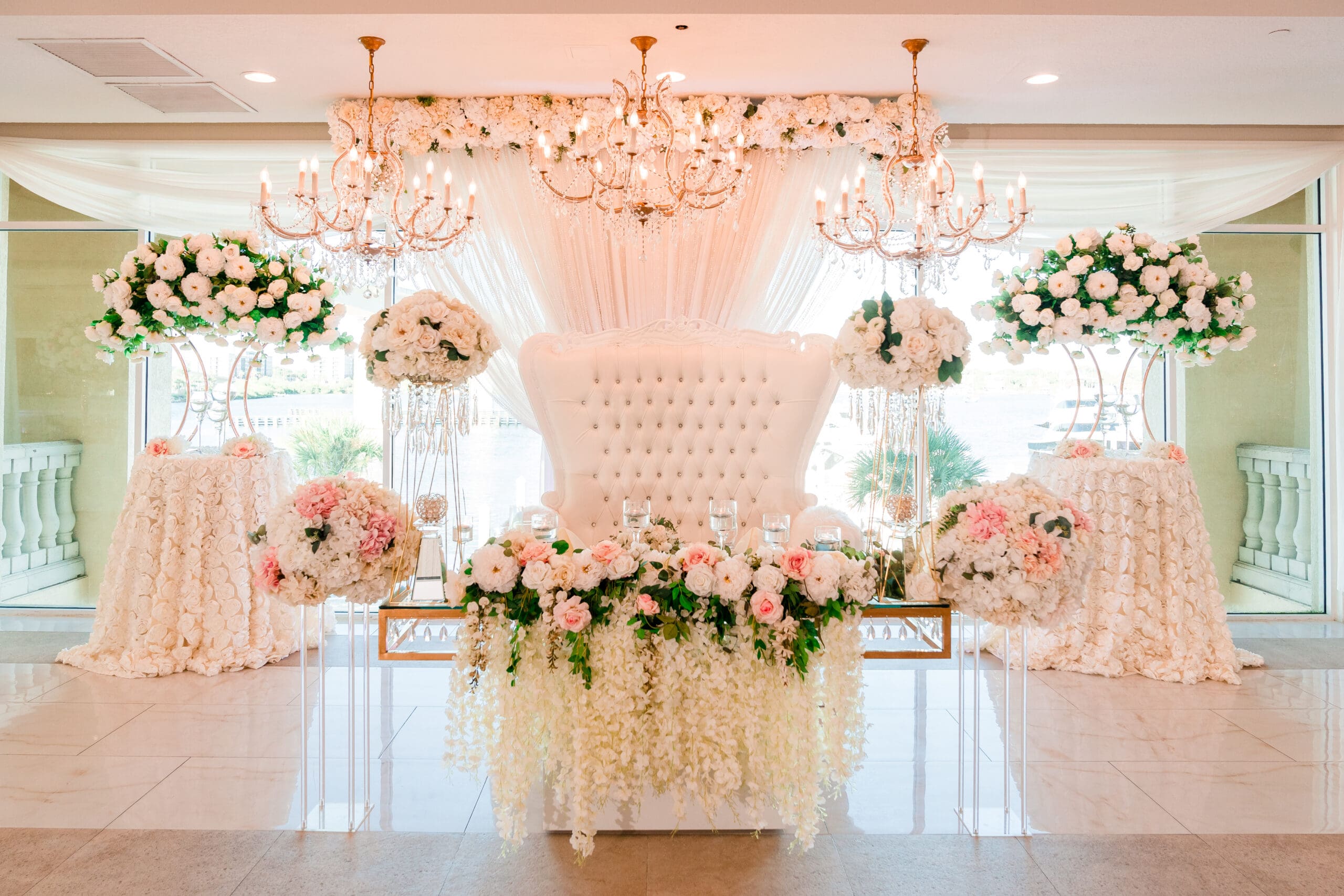 Jazer and Jen seated at a lavishly decorated table in the Crystal Ballroom reception area, adorned with white and pink rose bouquets, draped with white fabric, and featuring a white love seat.