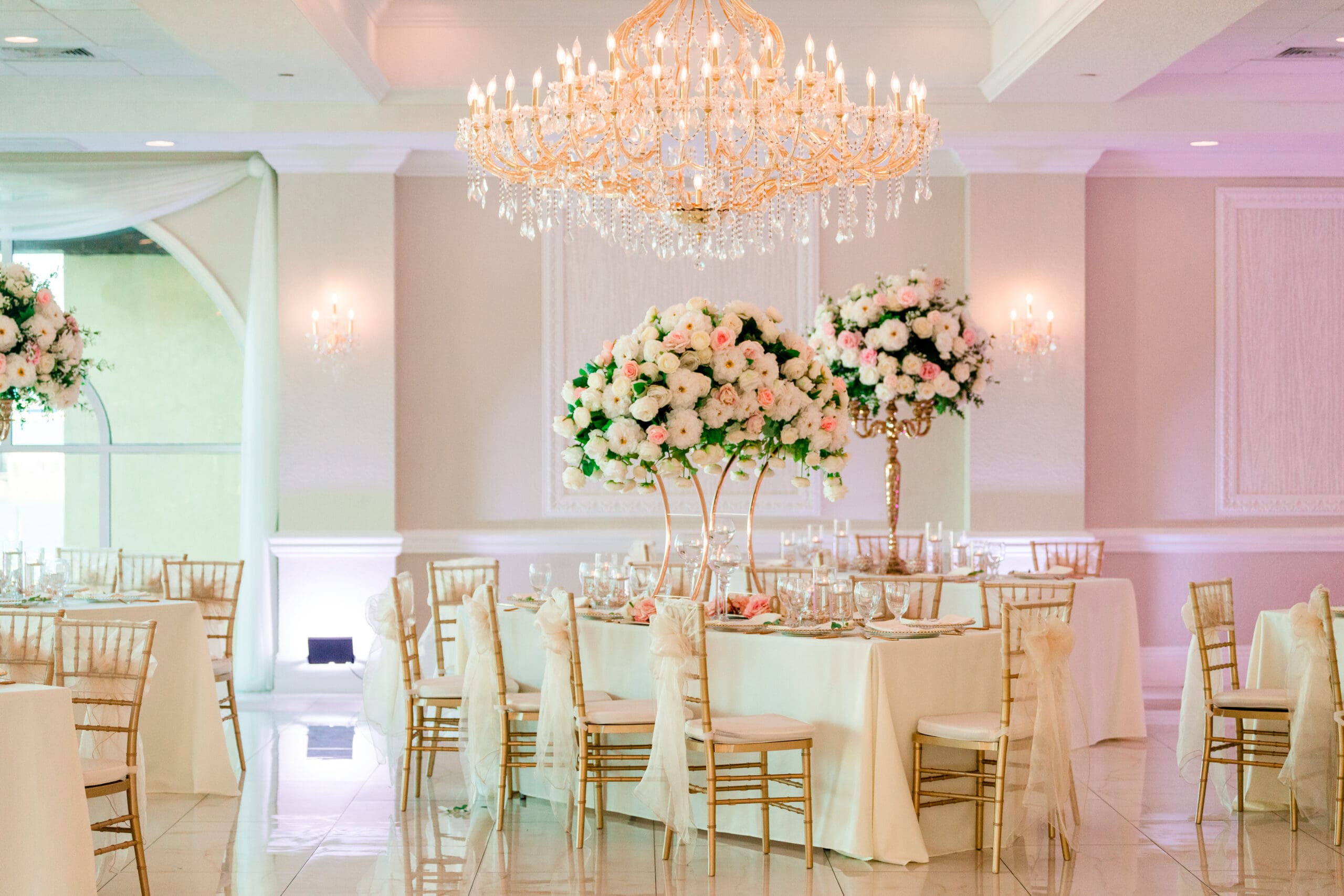 View of guests' tables adorned with large bouquets of white and pink flowers, white tablecloths, and chairs with white bows and veil-like drapes at the Crystal Ballroom.