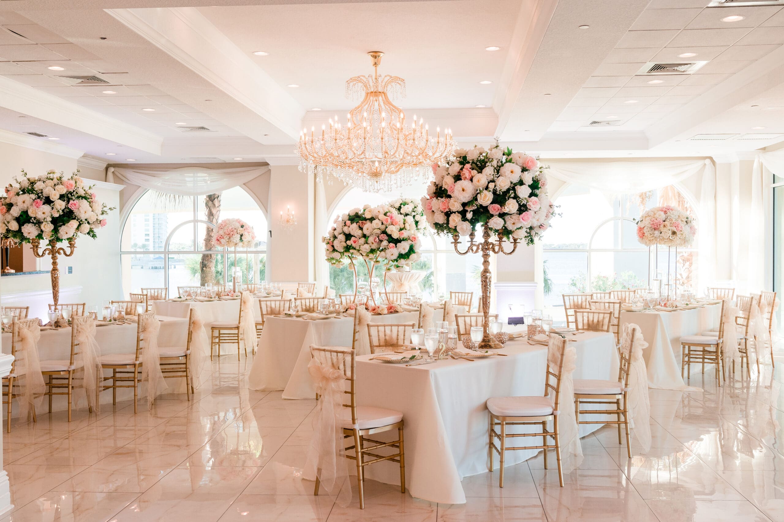 Wide shot of the Crystal Ballroom at Sunset Harbor, showcasing guests' tables adorned with bouquets of white and pink flowers, white chairs with bows and veil-like drapes, large windows allowing natural light to fill the room.