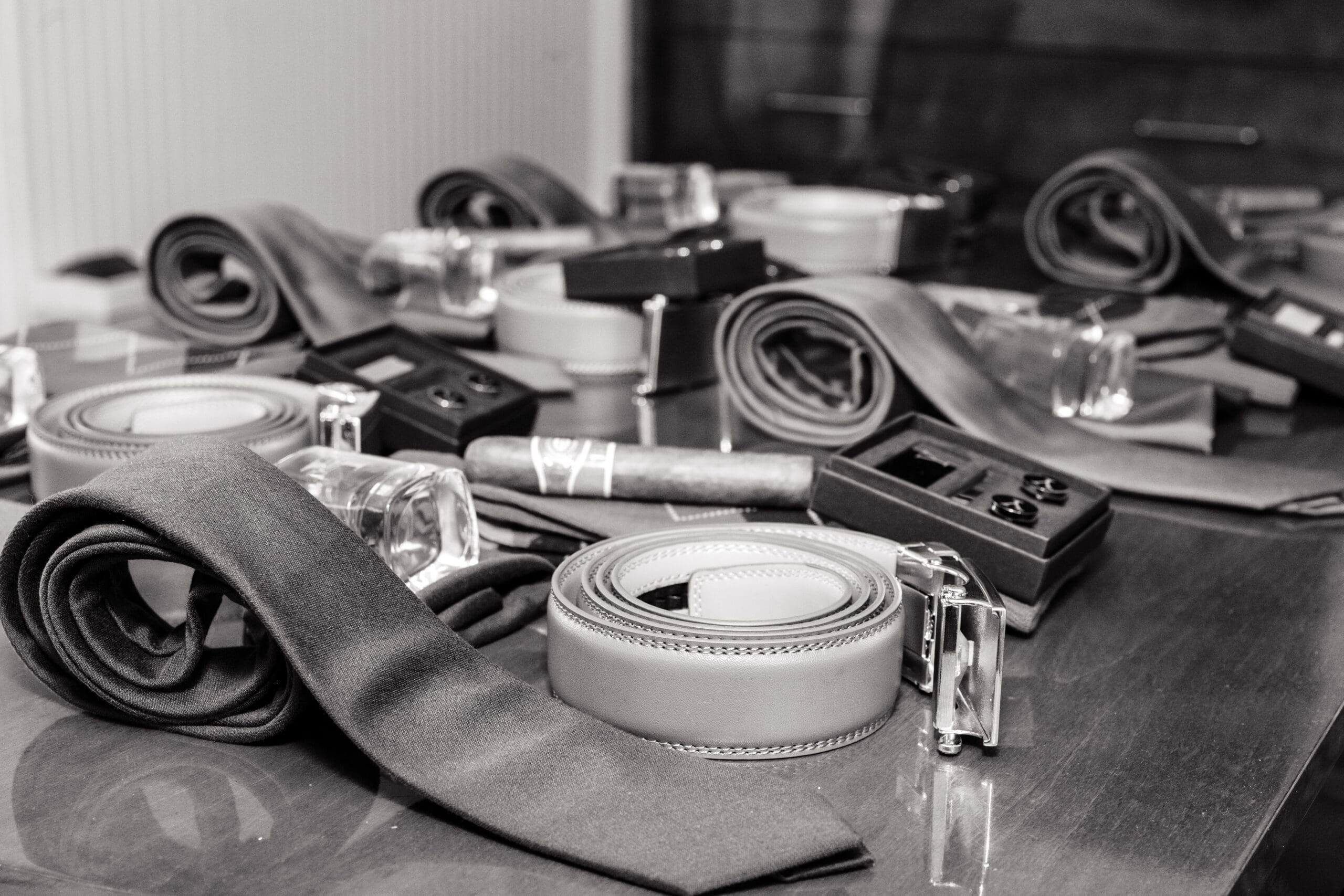 Black and white shot of groomsmen's matching ties, belts, cufflinks, cigars, and shot glasses laid out on a table.