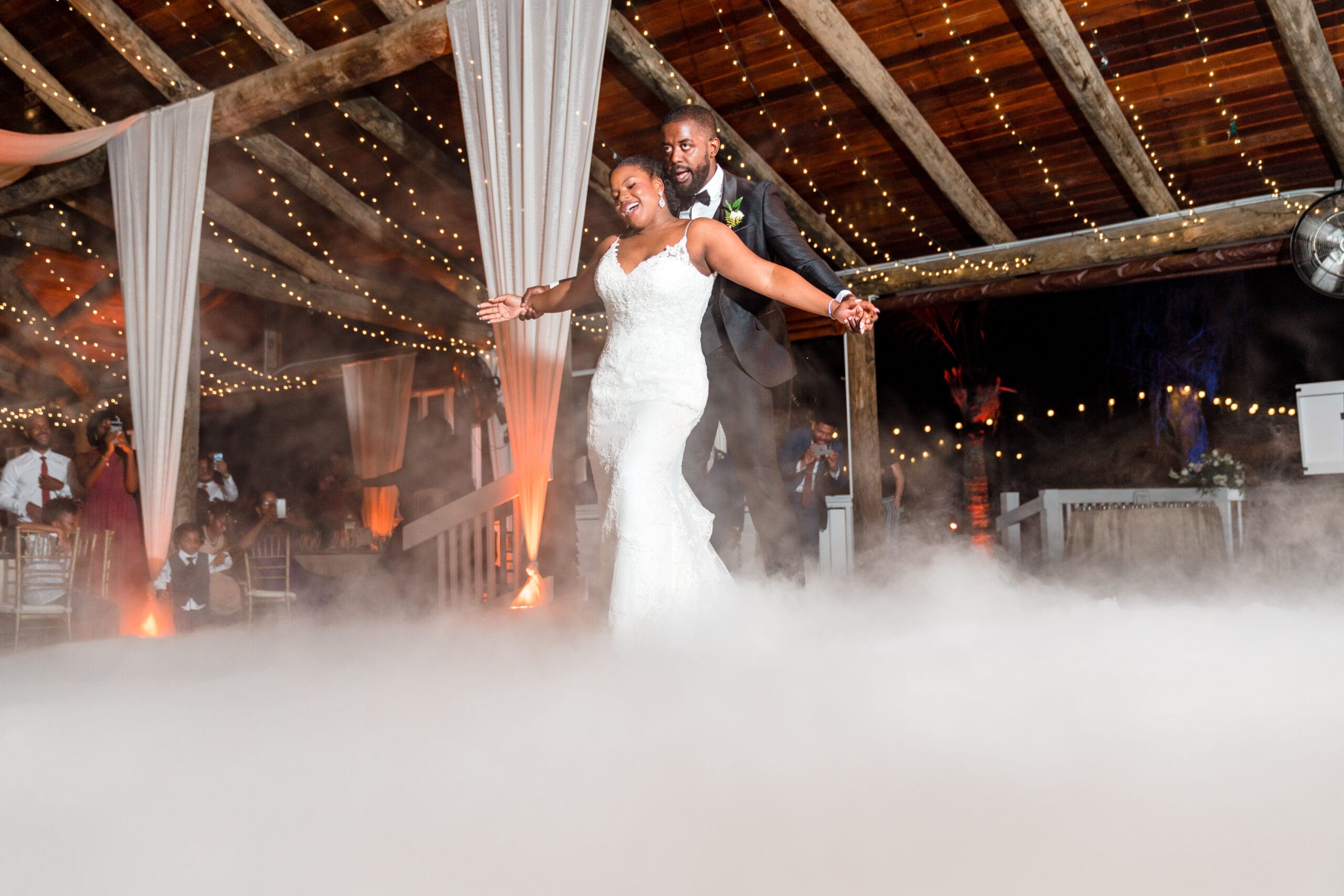 Wide shot capturing Jamie embracing Kourtni from behind with arms extended like wings, amidst the fog-filled dance floor at Paradise Cove reception.