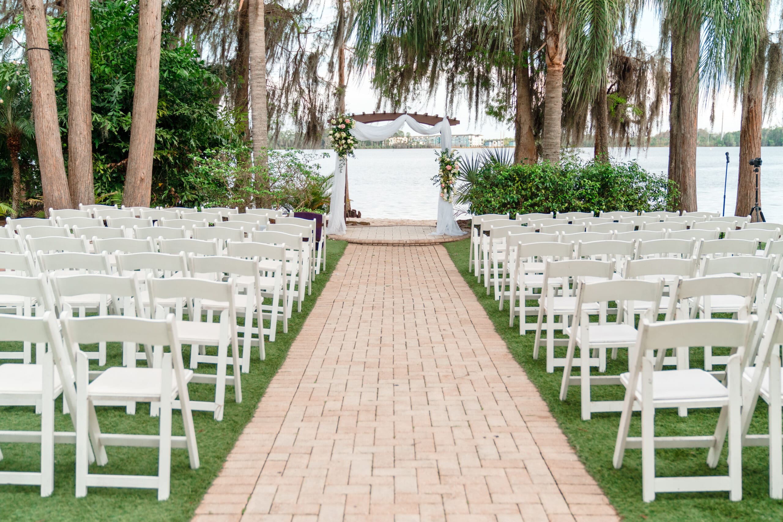 Empty outdoor ceremony area at Paradise Cove Wedding, showcasing white chairs for guests, an elegant altar draped with white drapes and adorned with flowers, with palm trees on each side, and clear water in the background.