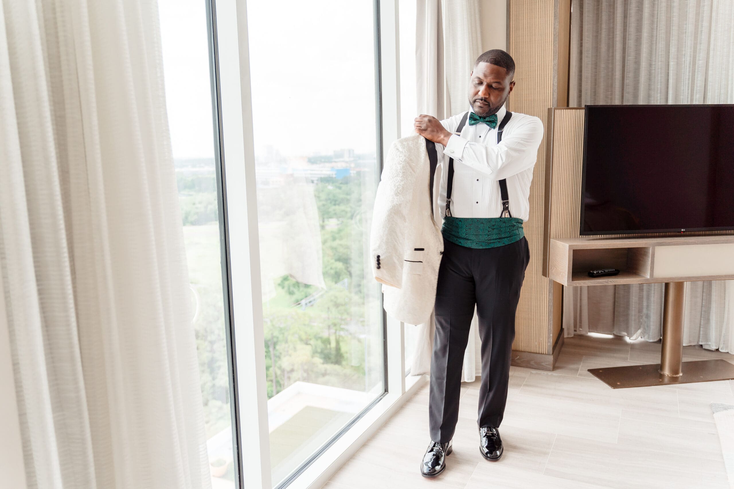 Glen stands by the window, donning his sports coat, in a hotel room with a picturesque view.