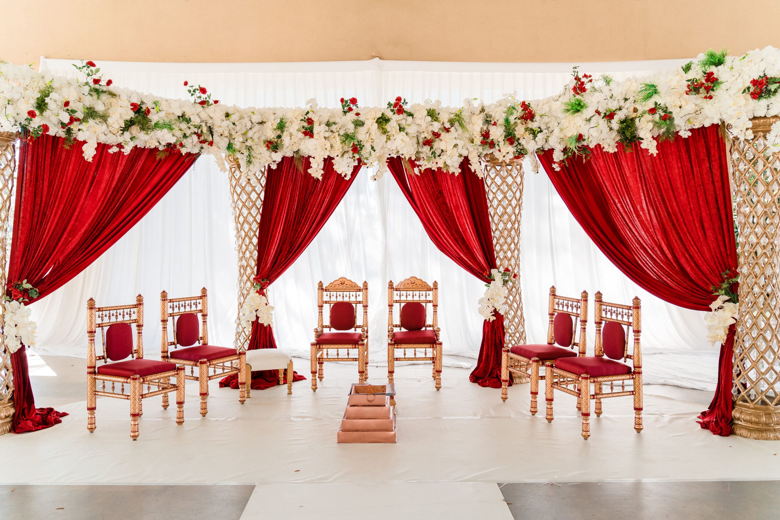 Traditional Indian wedding altar with red and gold decorative chairs, red drapes, and white flowers