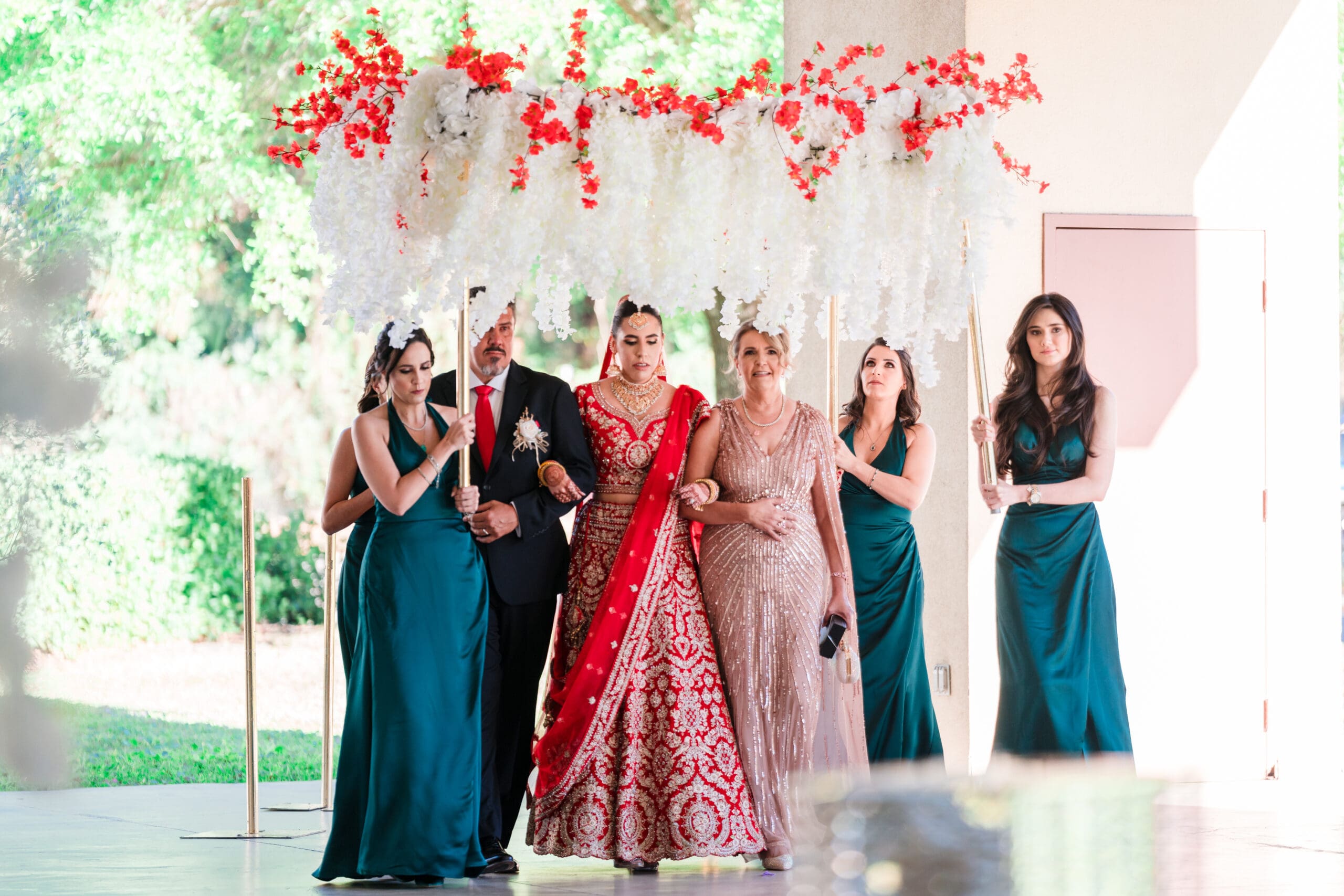 Sabrina walking with her parents and bridesmaids under a canopy of flowers at the traditional Indian wedding ceremony at Kraft Azalea Gardens