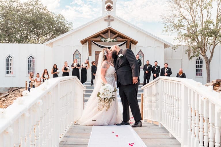 Newlyweds' Passionate Kiss on Bridge with Wedding Party and Church in Background at Hidden Barn