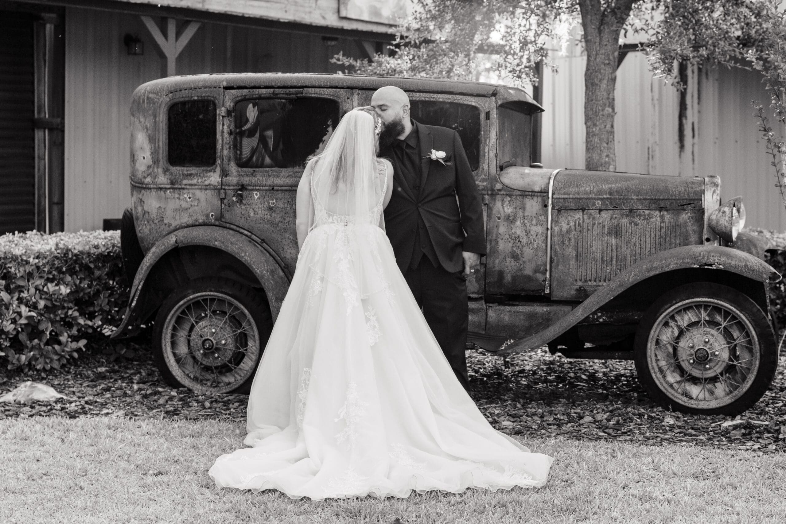 Newlyweds Kissing in Front of Rustic Car at Hidden Barn Venue
