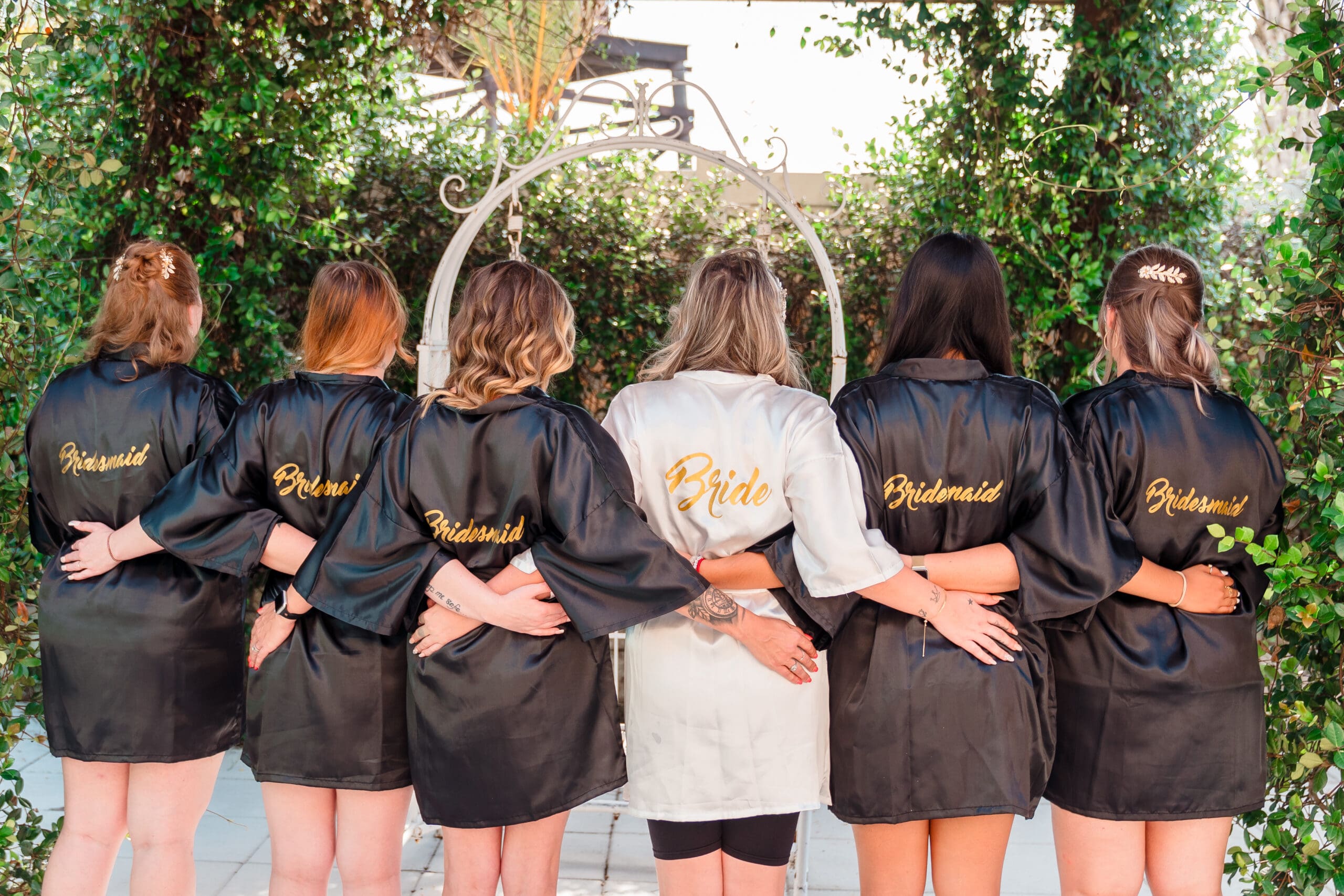 Taylor and her bridesmaids wearing labeled robes, Taylor's robe is white with "Bride" in gold, others in black with "Bridesmaid" in gold, arms intertwined, facing away from the camera