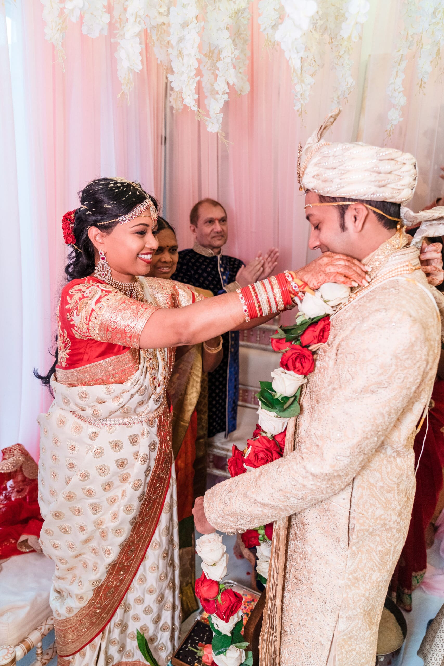 A traditional Indian couple, the bride adjusting her husband's collar, both dressed in exquisite traditional attire.
