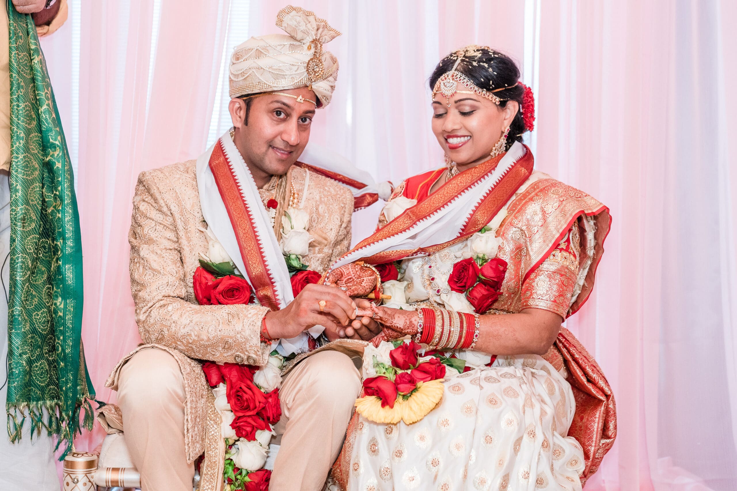 Smiling couple in traditional Indian wedding attire, the husband placing a ring on his bride's hand during the ceremony.