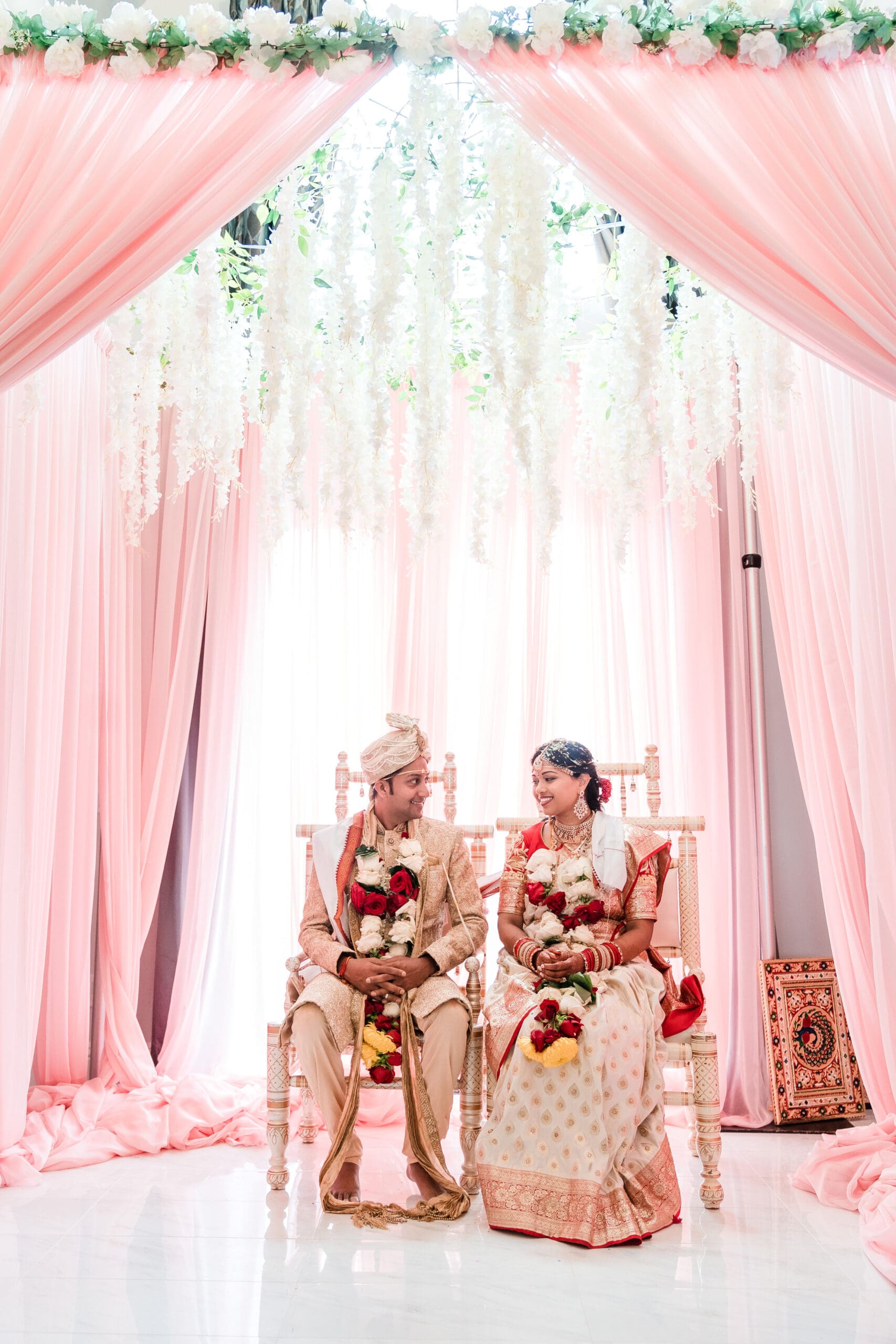 Couple in traditional Indian wedding attire holding flowers, sitting in front of window with pink decorative drapes - cultural wedding photography