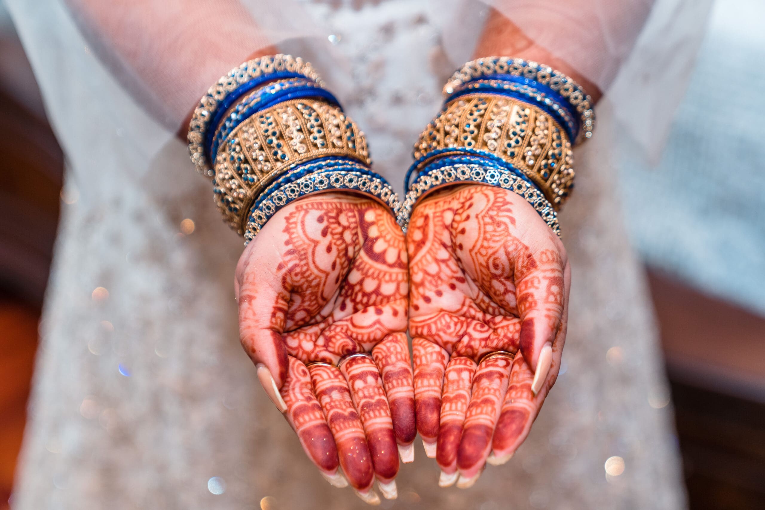 Close-up of bride's hands adorned with gold and blue bracelets, showcasing intricate henna tattoos, with a blurred white dress in the background.