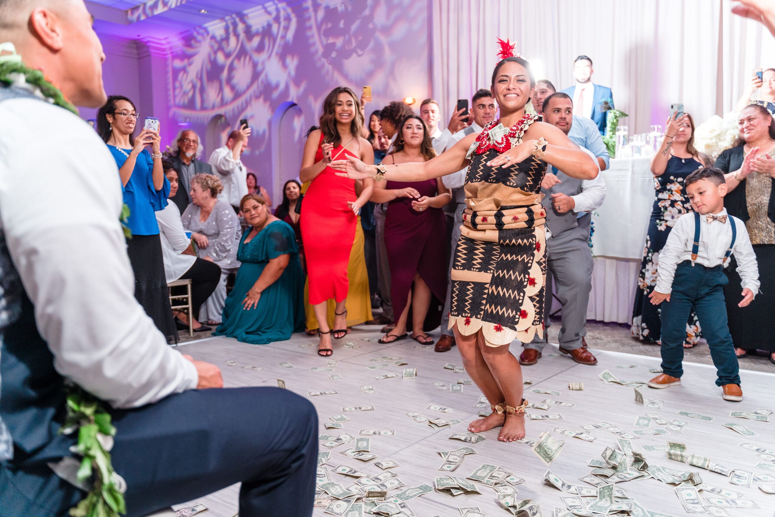 Bride dancing joyfully in traditional Hawaiian attire amidst scattered money on the ground, performing a lively hula move with a radiant smile, surrounded by friends and family dancing in celebration.