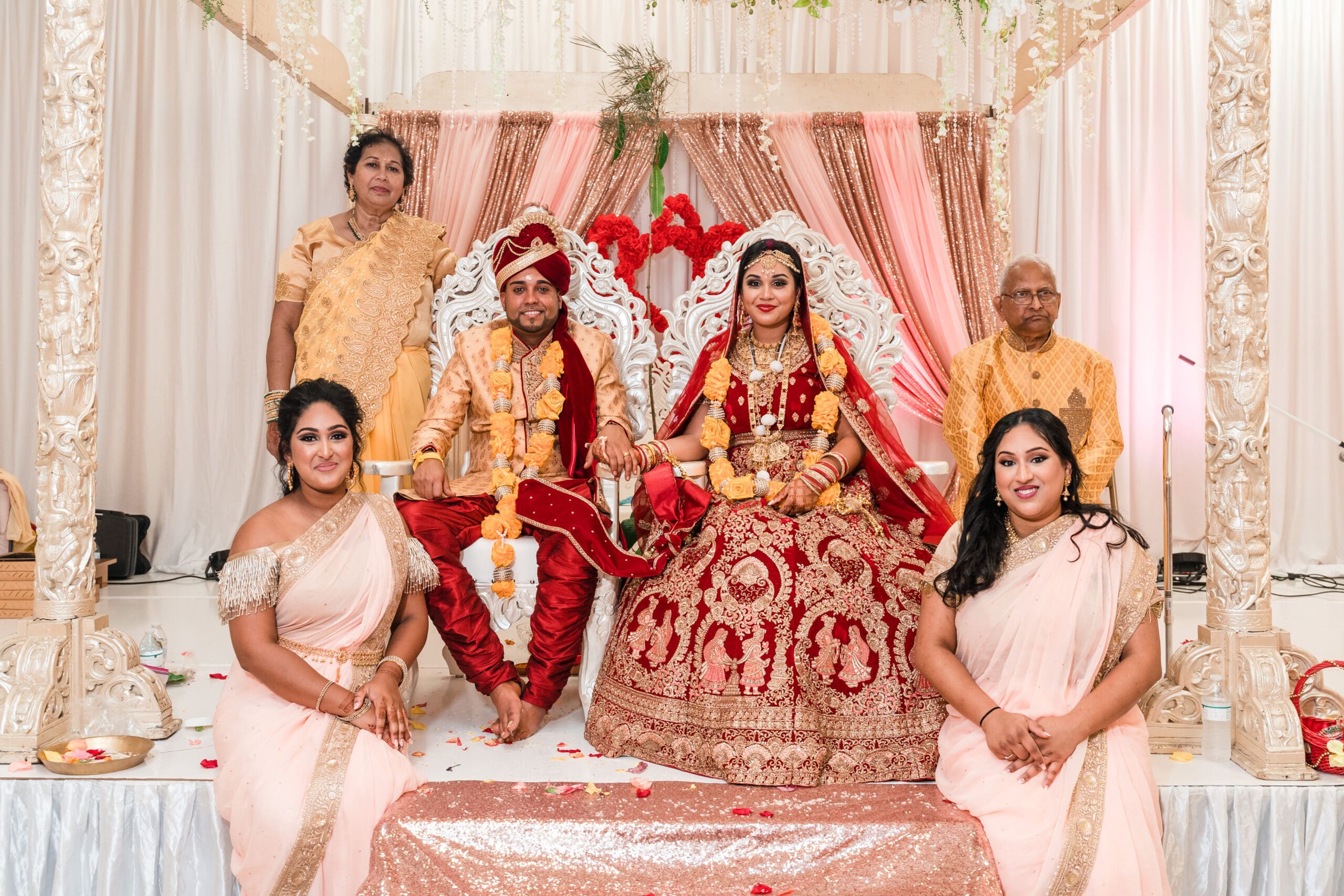 Bride, groom, bride's parents, and two sisters seated together, all dressed in traditional Indian wedding attire, smiling in cultural wedding portrait.