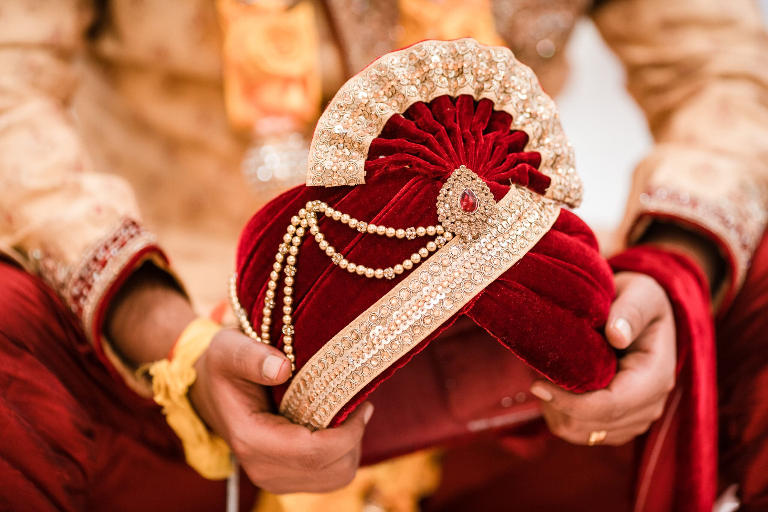 Traditional red headpiece adorned with gold, worn by the groom as part of an Indian wedding attire.