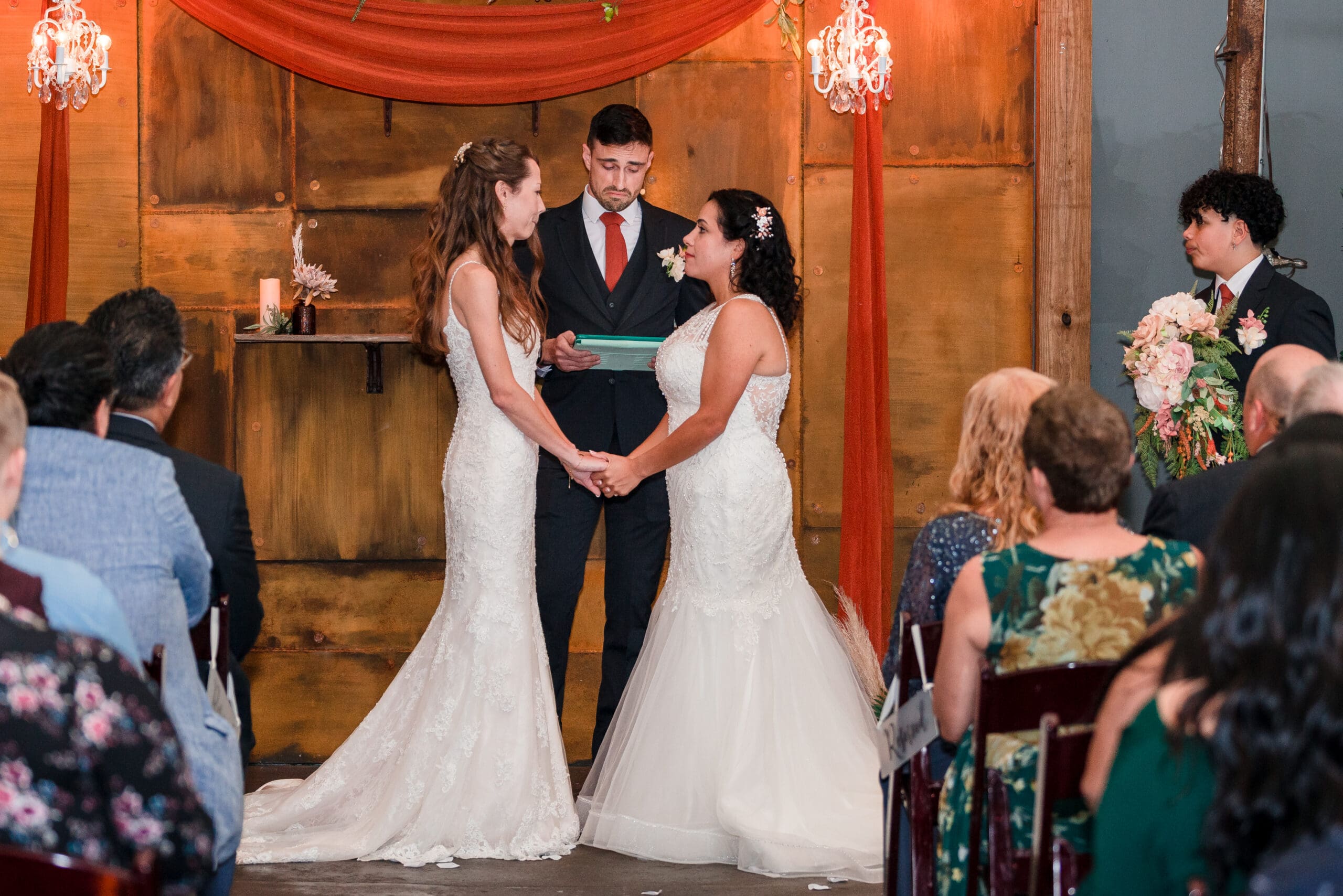 Two brides holding hands and looking into each other's eyes at the altar, surrounded by family and friends, with the officiant visibly moved by the love shared between them