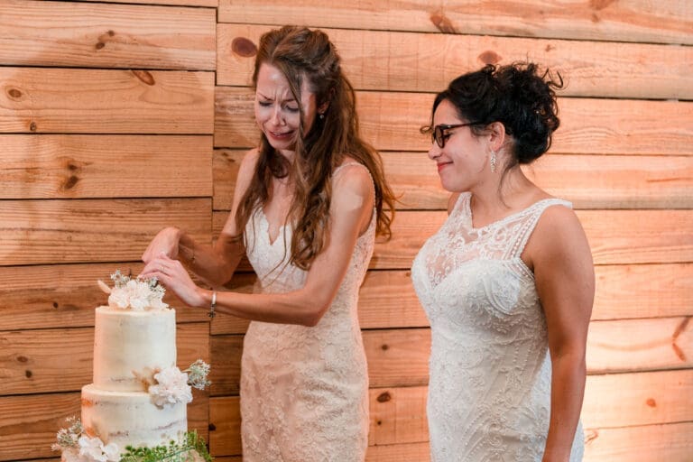 Emotional moment as one bride tears up while cutting wedding cake with her partner, both dressed in white.