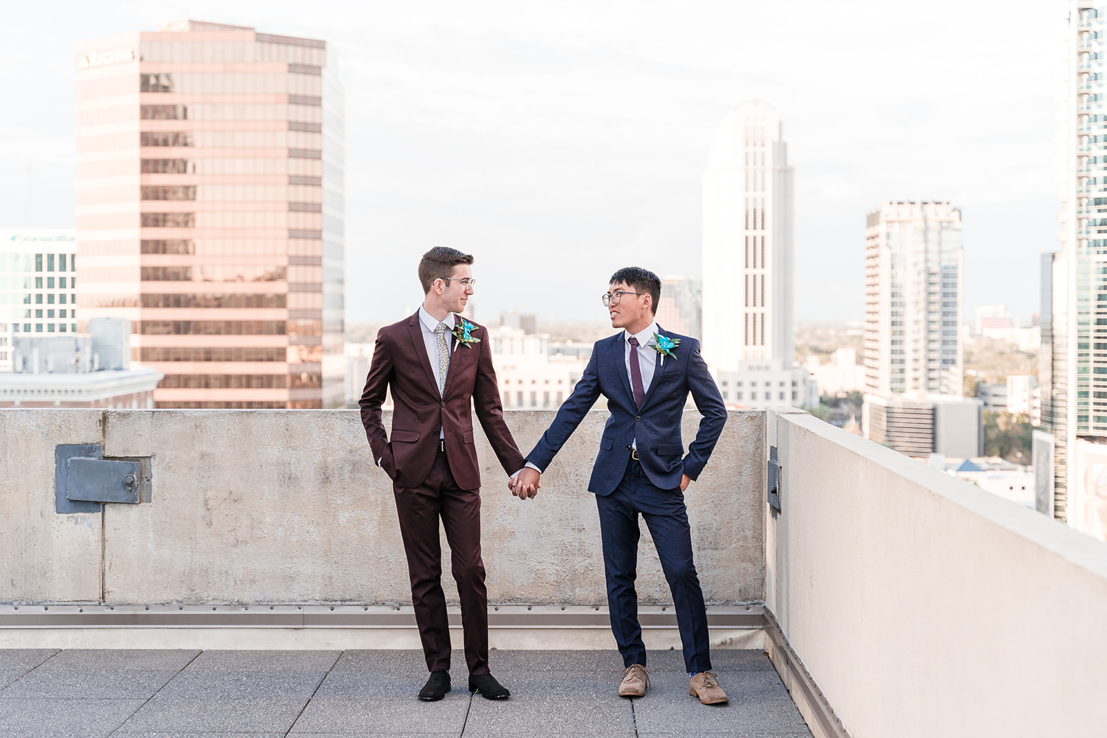 Two grooms take a picture, holding hands on the rooftop of a building in downtown Orlando, FL, with the beautiful cityscape as the backdrop.