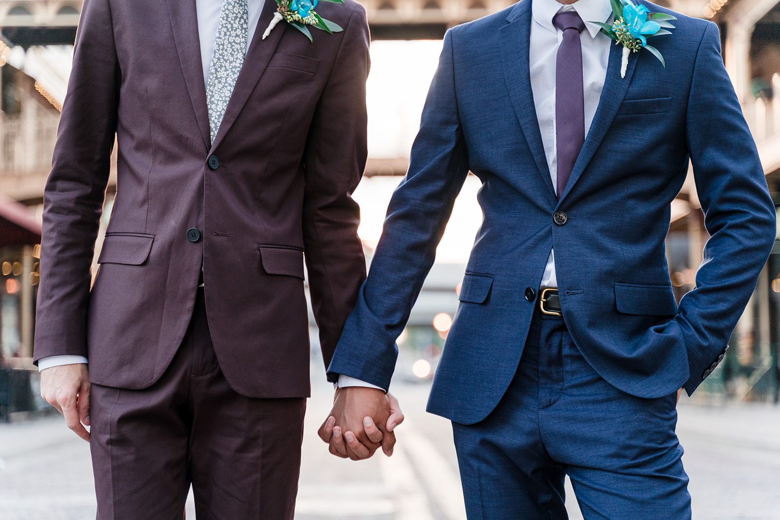 Two grooms hold hands, one wearing a stylish blue suit and the other in a dashing purple suit, symbolizing their unique personalities and shared love.