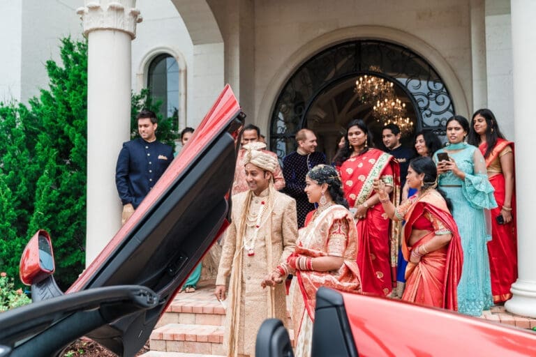 Bride and groom exit in style in a McLaren, dressed in traditional Indian wedding attire, with the wedding party in the background also in traditional attire.