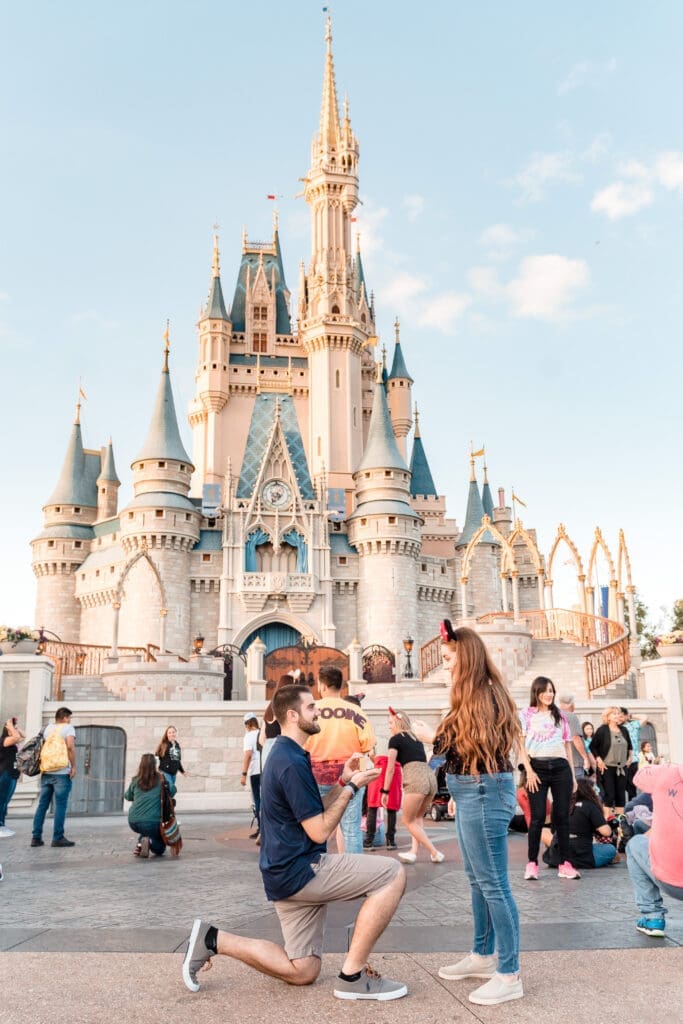 Enchanting proposal at Magic Kingdom: Capturing the magical moment in front of the main castle, with her wearing Disney ears, adding authentic Disney charm to the proposal filled with love and Disney cheer on a beautiful day out.