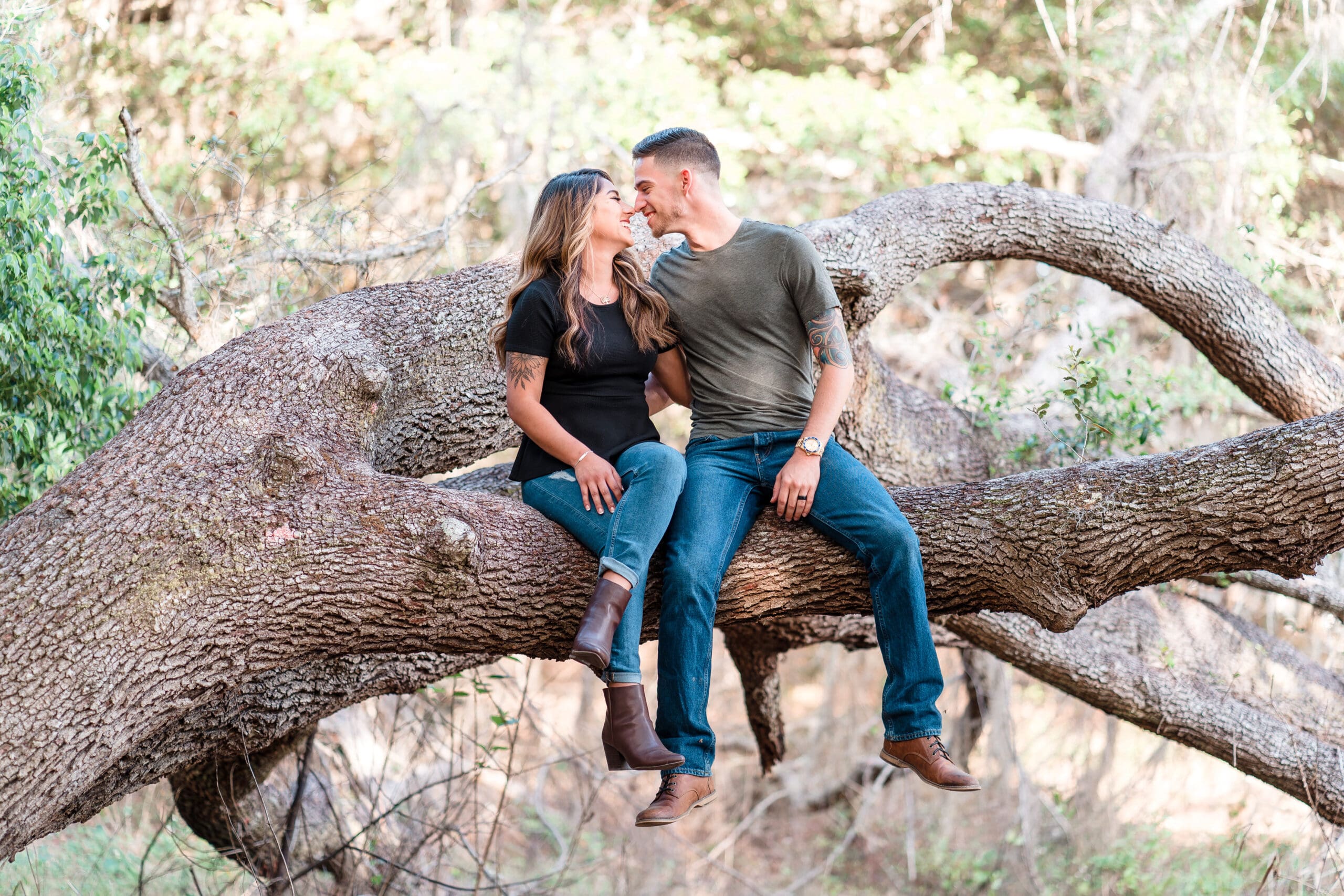 Engagement photo of couple sitting on a tree branch, touching noses, looking into each other's eyes with nature in the background - very unique picture.