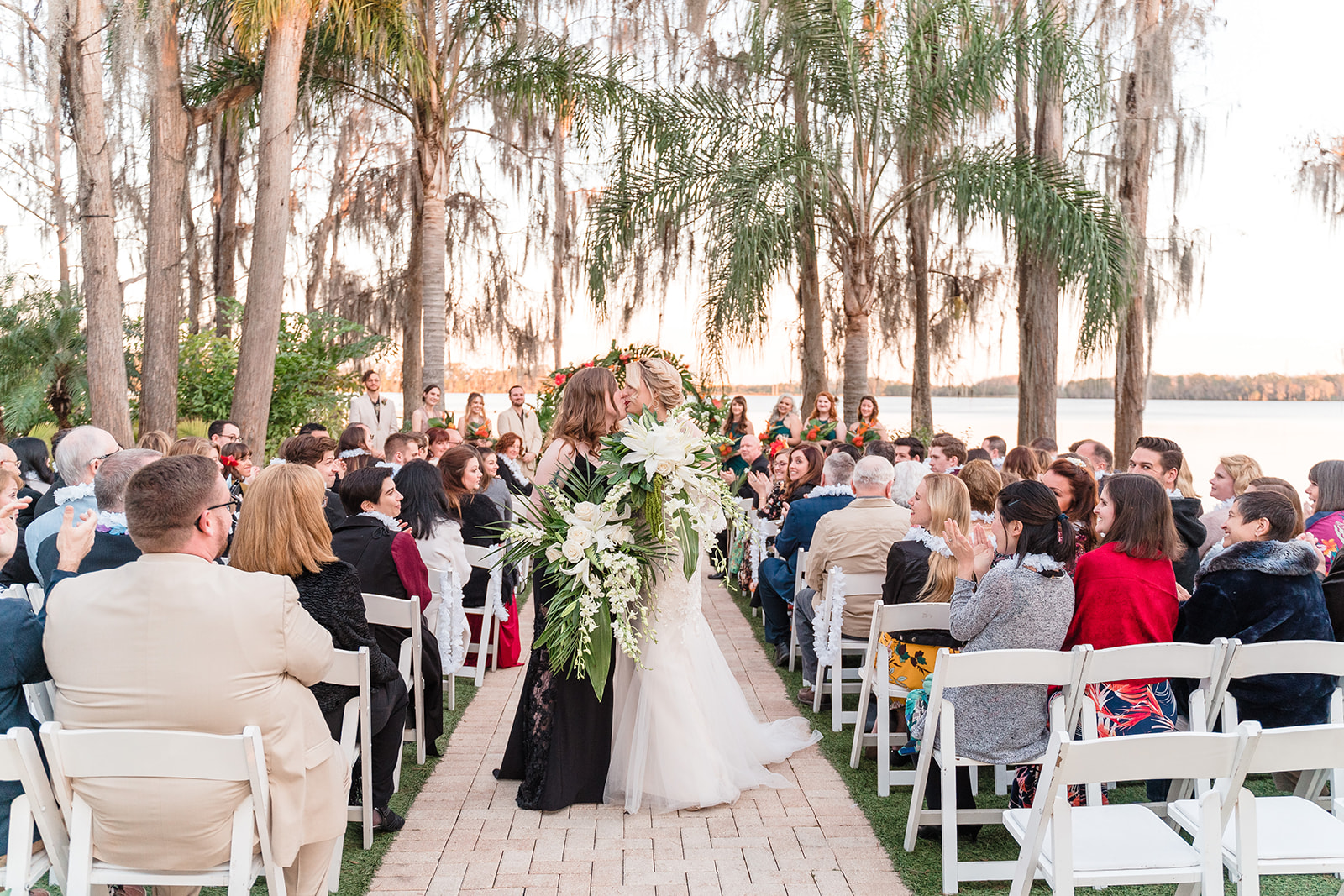 Two brides share a kiss as they walk down the aisle together, surrounded by smiling and applauding guests, with a backdrop of lush palm trees and a serene Florida lake.