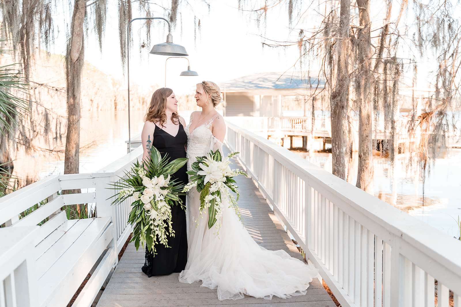Two brides standing on a bridge, gazing into each other's eyes with love and affection, both holding flower bouquets, one in a black dress and the other in a white dress.