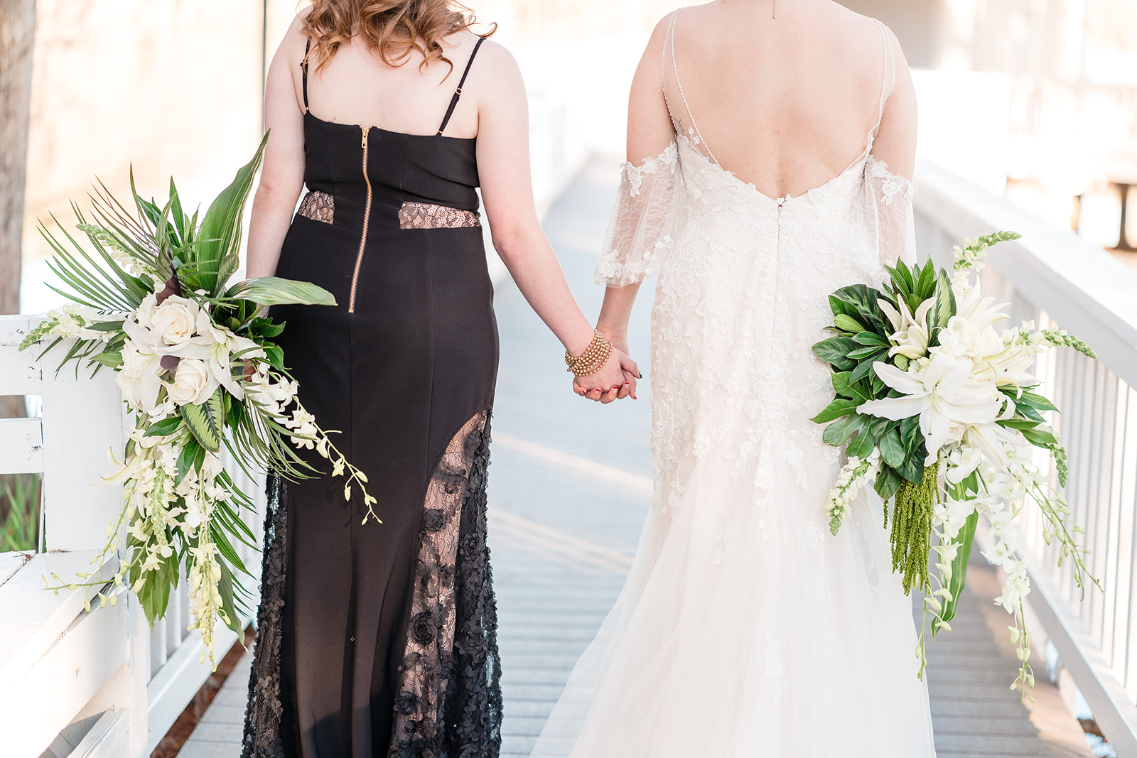 A close-up of two brides holding hands on a bridge, each with a bouquet of flowers, one in a black dress and the other in a white dress.