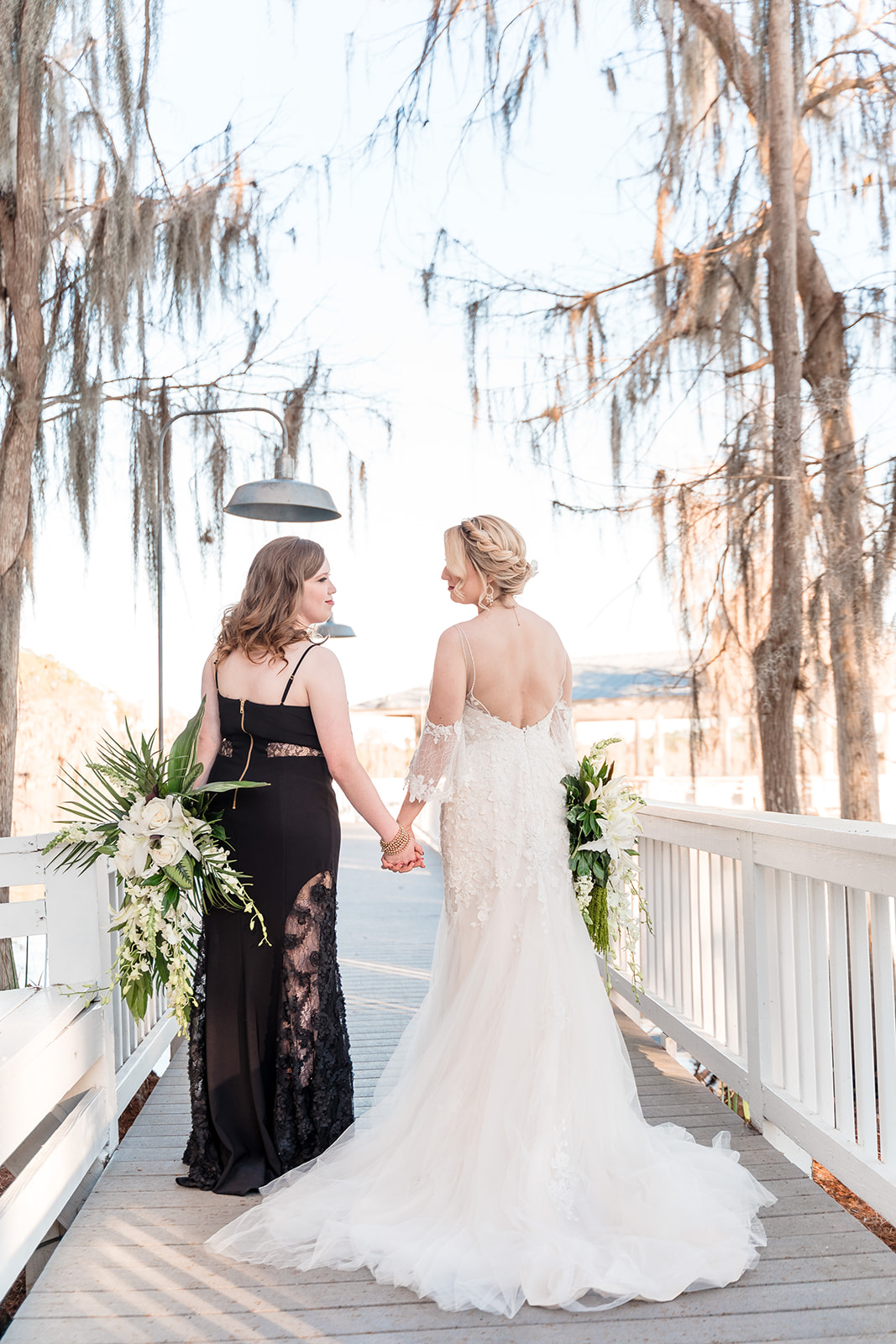 Two brides holding hands on a bridge, one in a black dress and the other in a white dress, symbolizing their union in love and harmony.