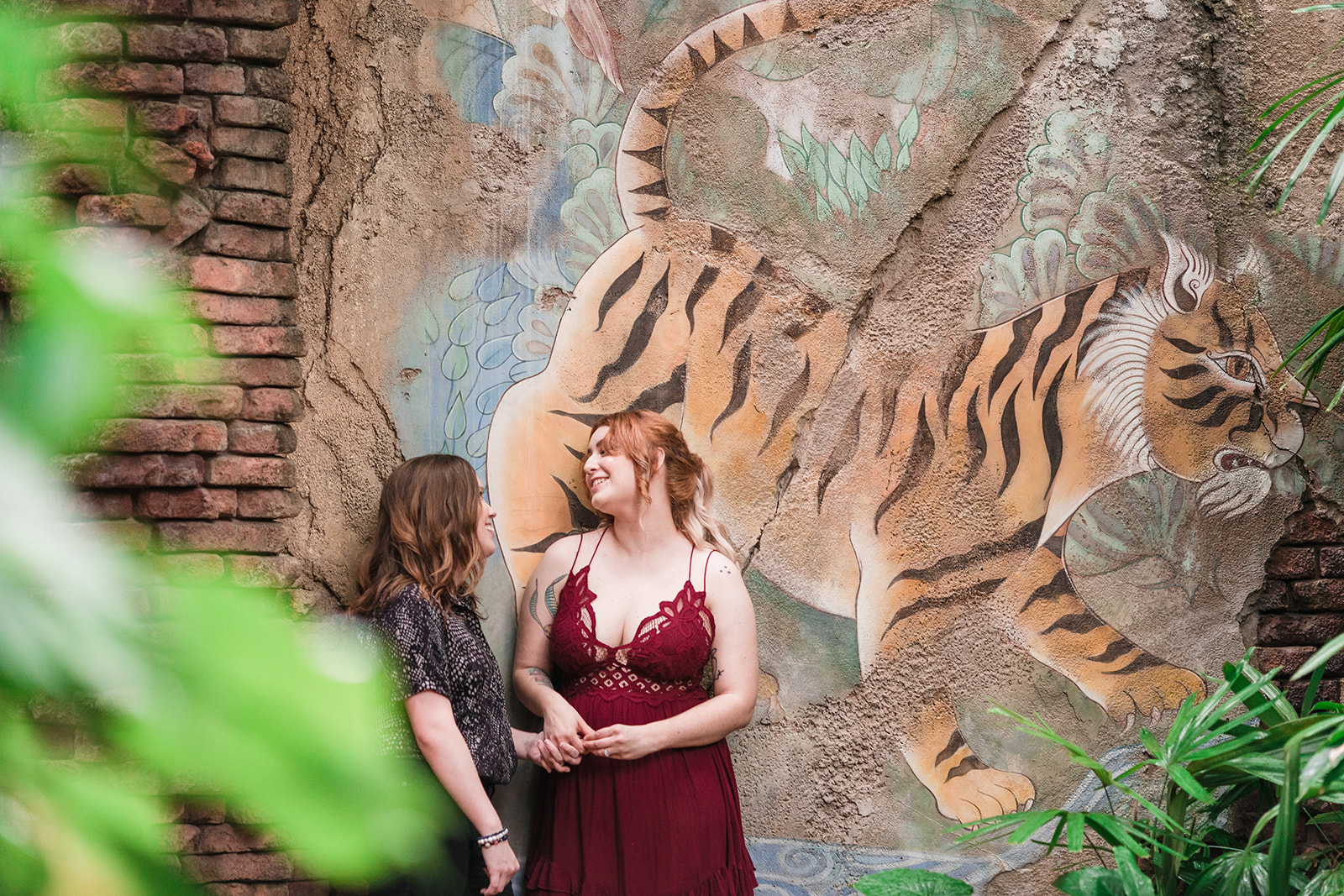 Amidst a wall painting of a tiger, two brides embrace, one kissing the other's cheek, while the vibrant scenery adds to the enchantment of their love.