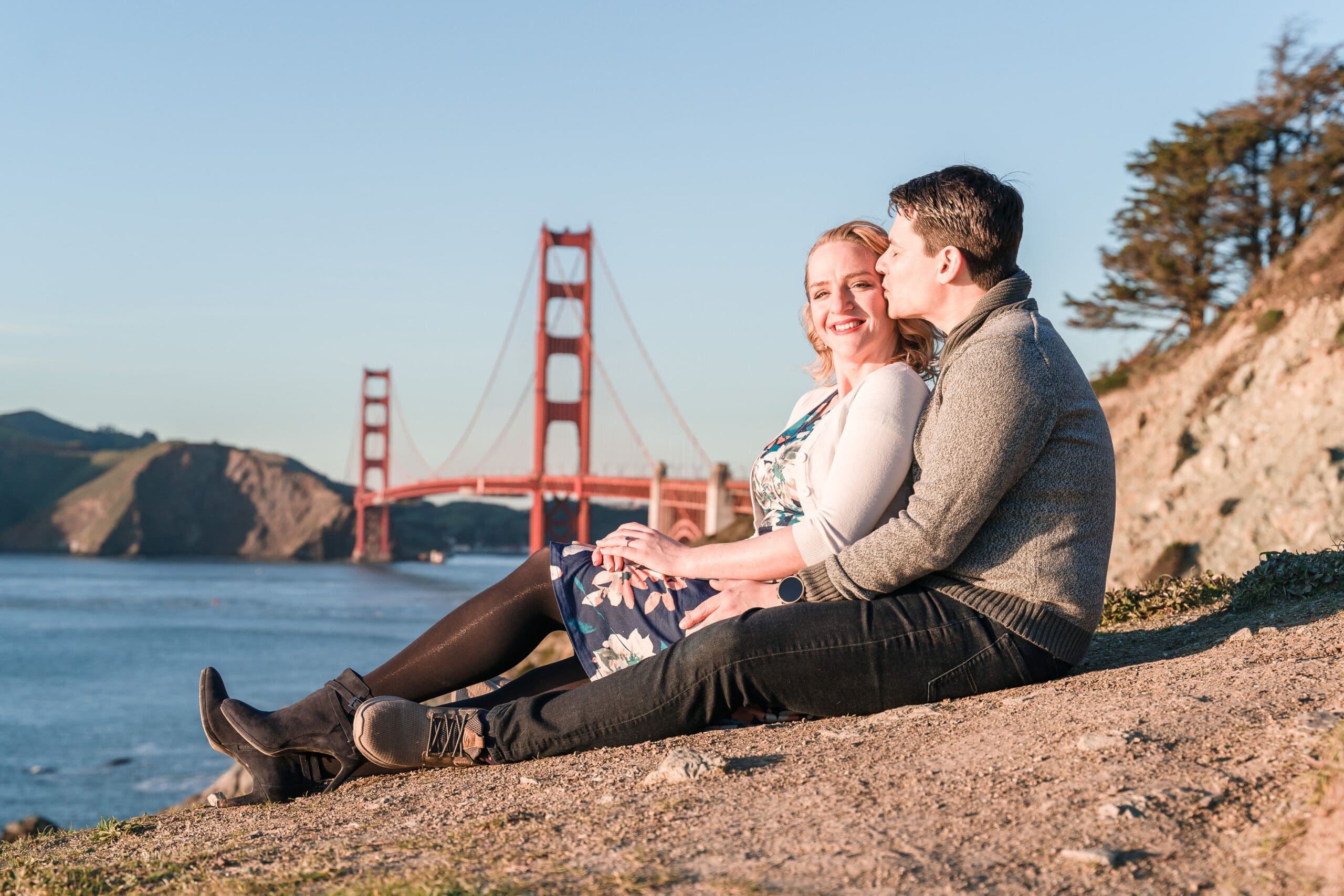 Romantic moment overlooking San Francisco: Couple shares an intimate embrace as the soon-to-be wife sits in the lap of her future husband on a cliff, with the iconic San Francisco bridge in the background.