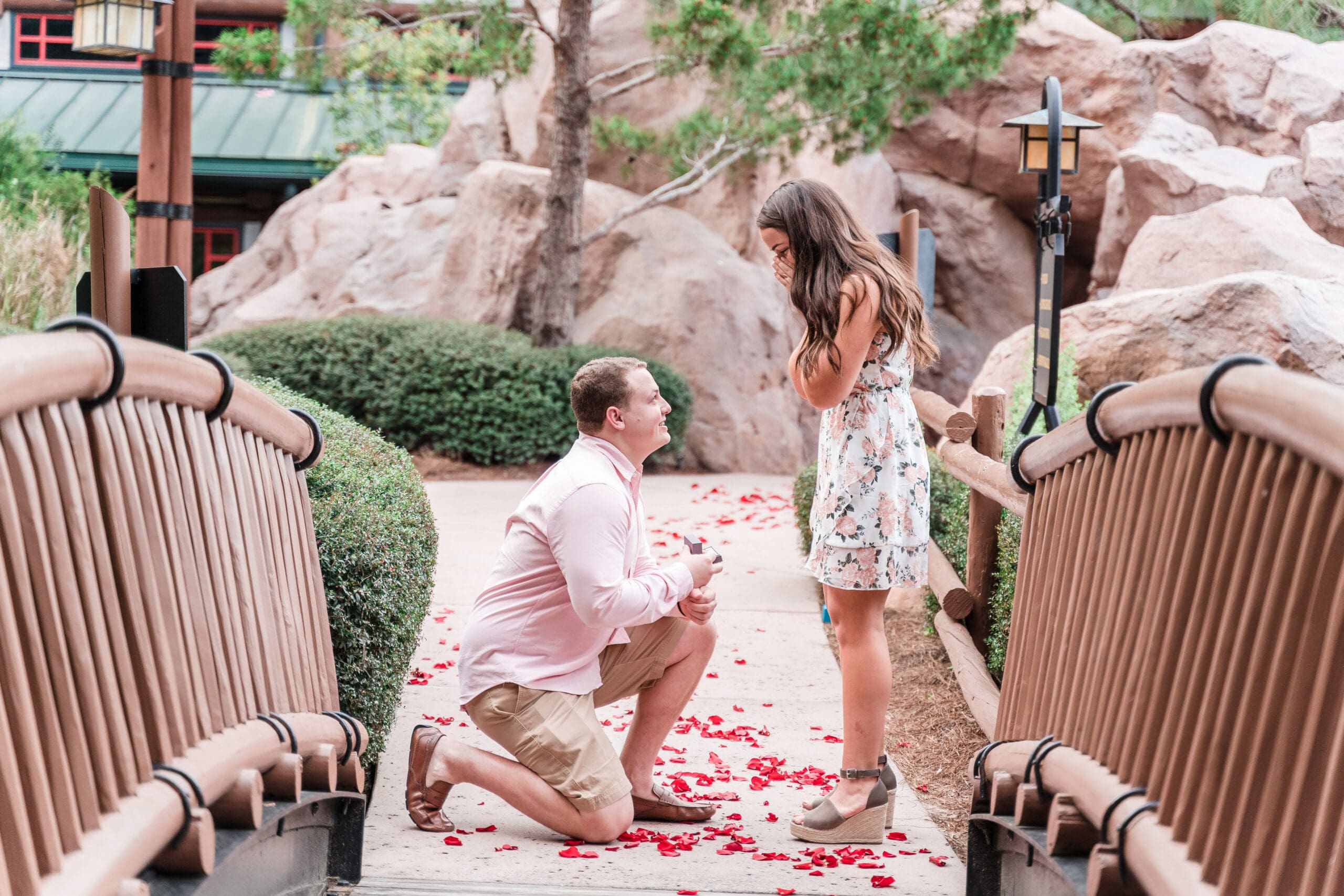 Magical proposal at Disney Wilderness Lodge: Down on one knee as he asks the question, right in front of the bridge adorned with rose petals leading up to the walkway, capturing her gasp of surprise, showcasing the best engagement photo sessions in Orlando Florida with the team at Jerzy Nieves Photography.