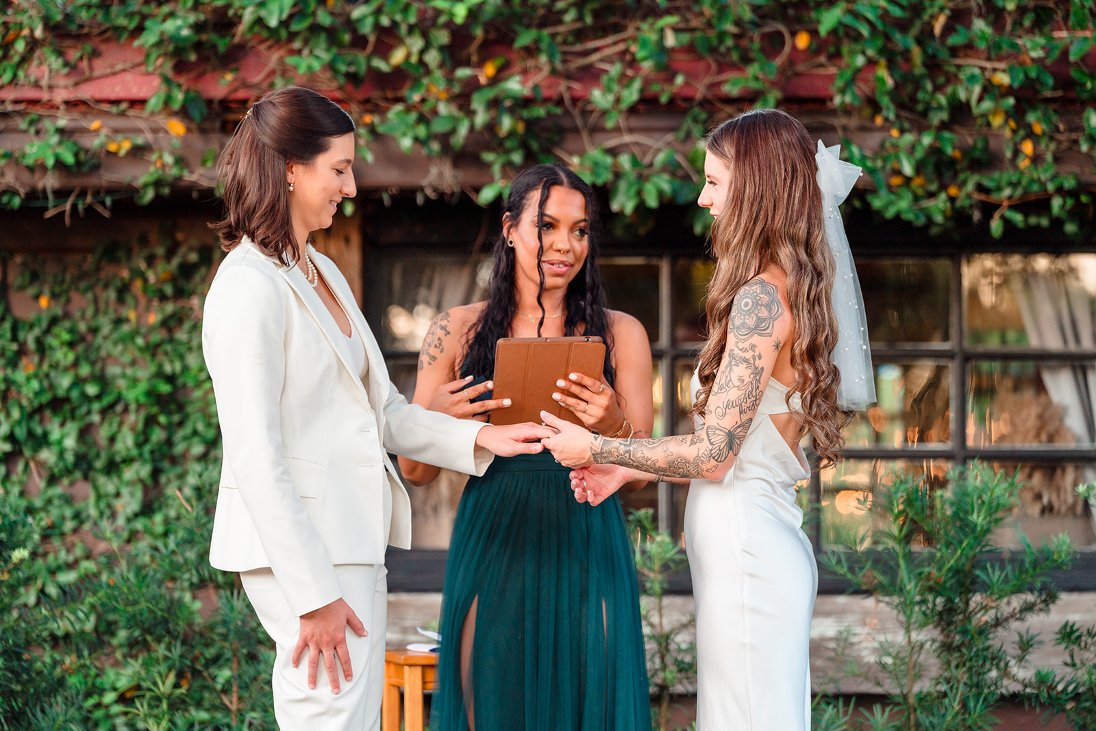Two brides holding hands at the altar, saying 'I do' with a female officiant holding an iPad, capturing the beauty of the background in this modern wedding.
