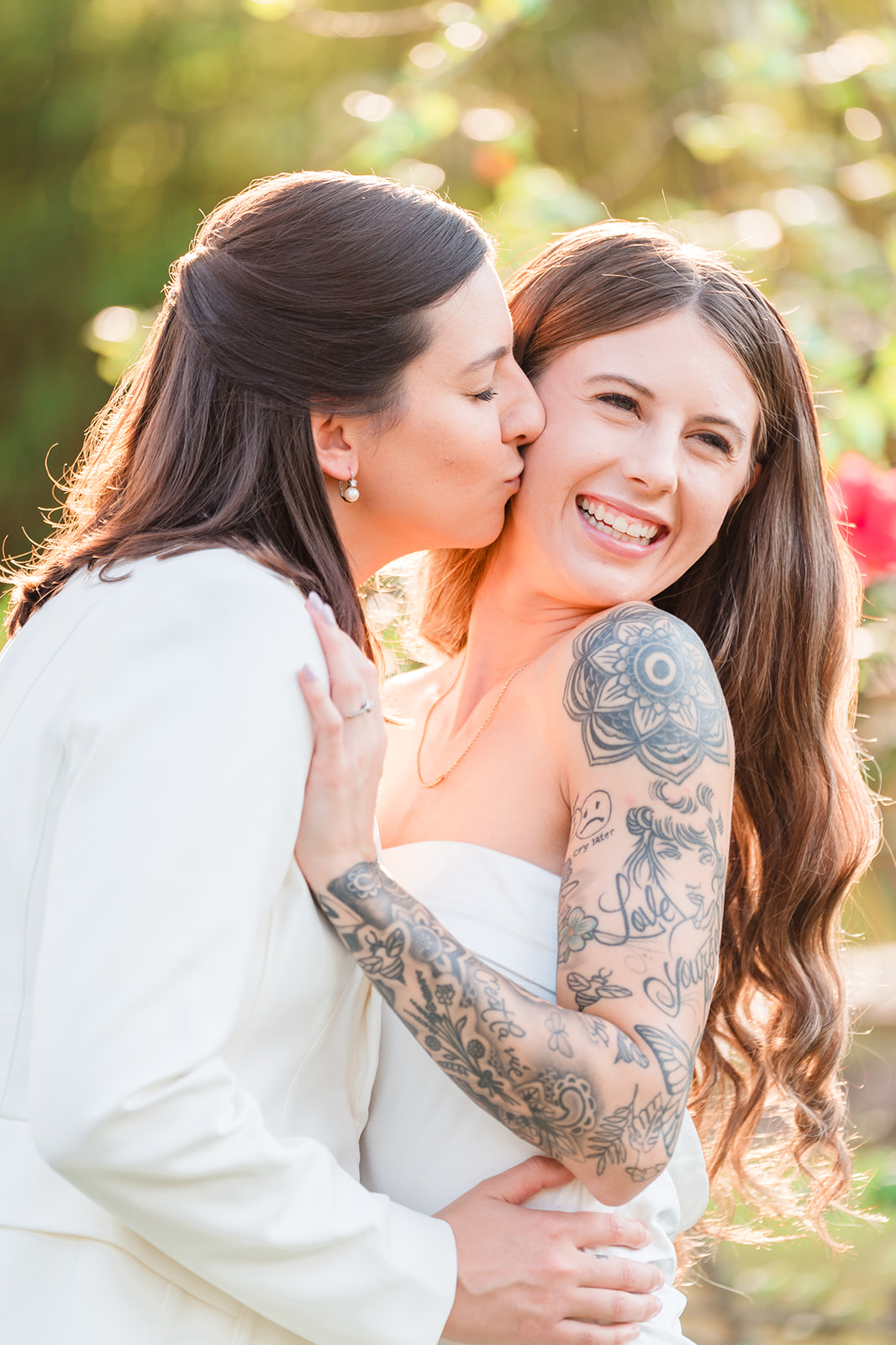 Two LGBTQ brides share a moment, one kissing the other's cheek, captured in perfect lighting.