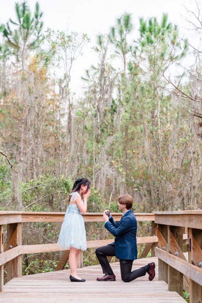 Scenic pier proposal: Man on one knee opening the ring case with nature in the background, while she clasps her mouth with both hands in surprise, captured by the best surprise proposal wedding company in Orlando FL, Jerzy Nieves Photography.