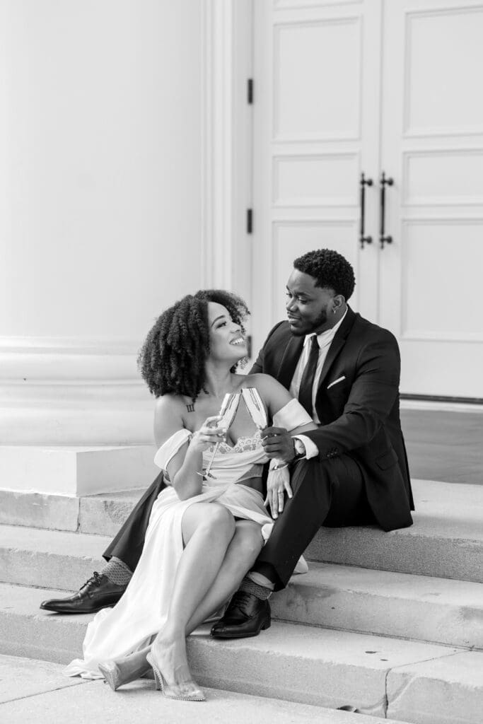 Black and white engagement photo of a couple toasting champagne on steps, captured in timeless elegance.