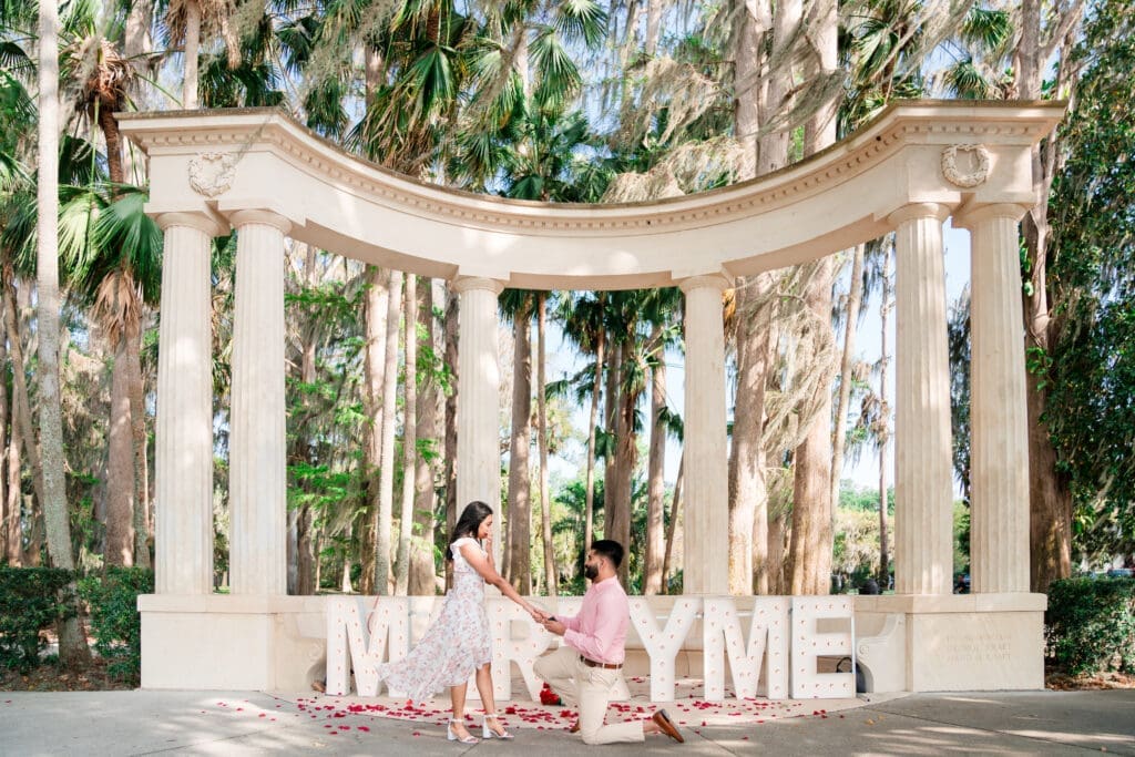 Mesmerizing proposal scene: 'Marry Me' sign in the background, adorned with Roman-style pillars and rose pillars on the floor, capturing the moment of surprise as she gasps with her hand on her mouth when he asks the question.