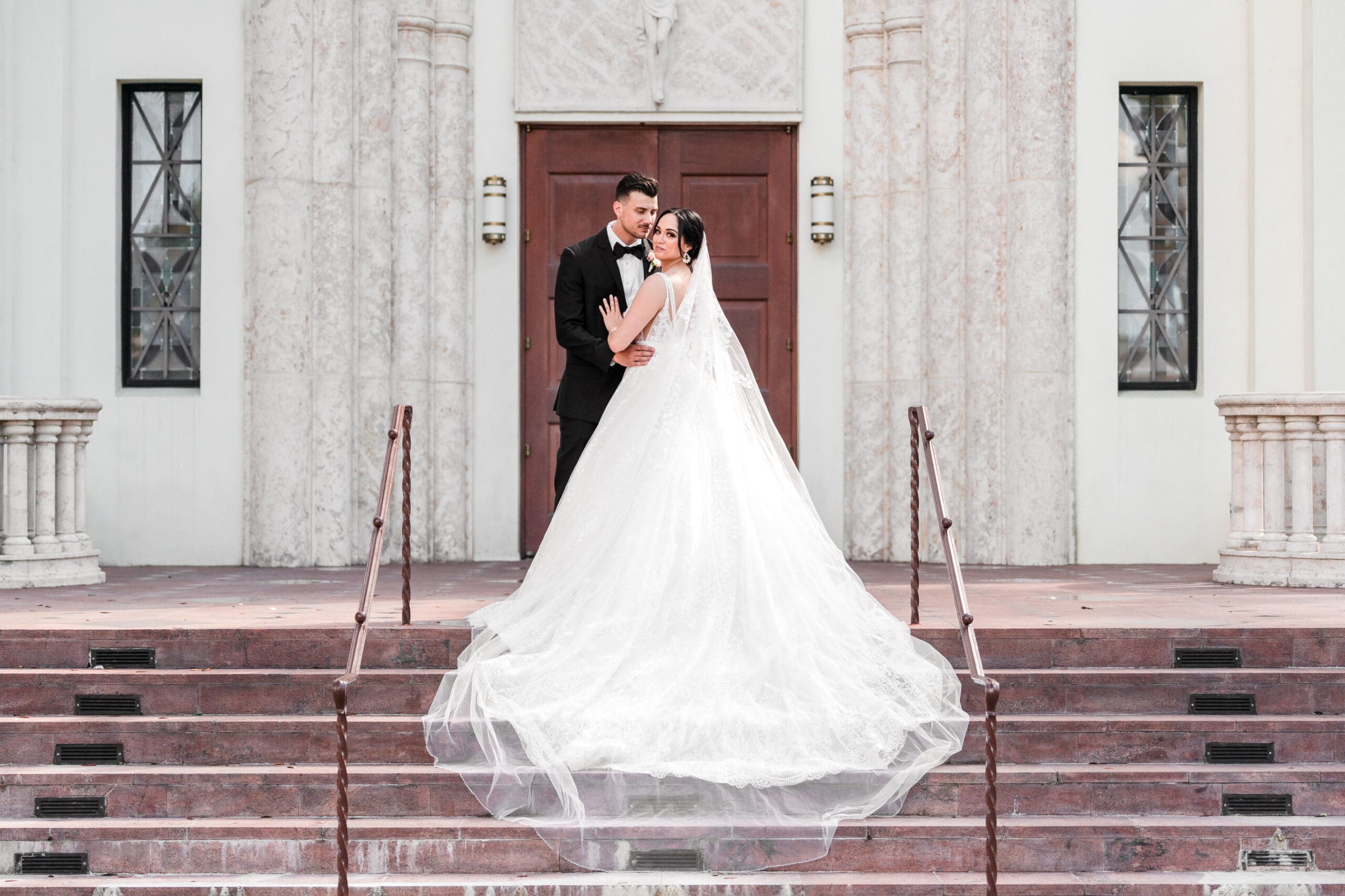 Couple standing on the stairs of a church, with the bride's dress flowing down the steps as they stand at the top, the bride looking at the camera while the husband admires her.