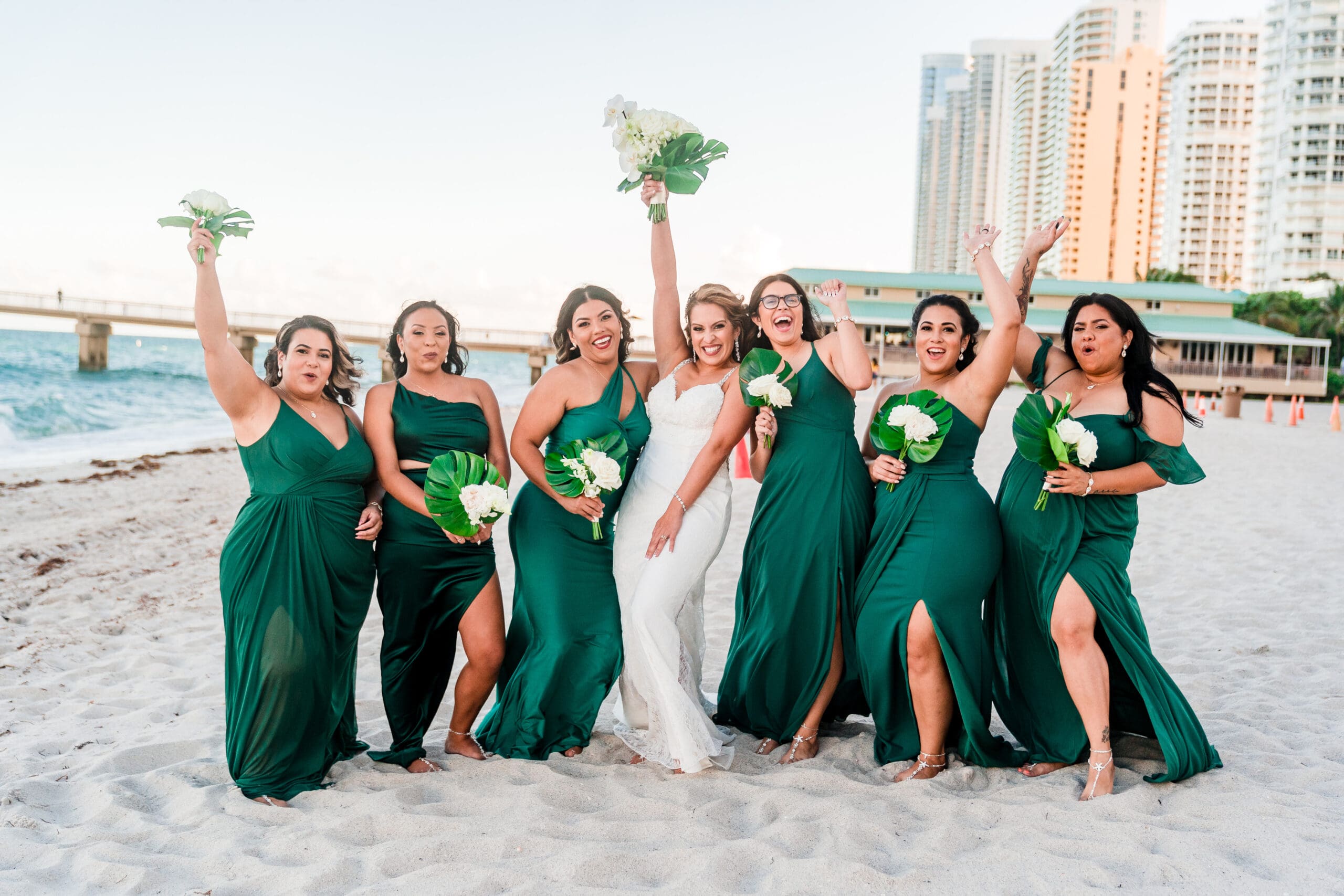 Beach wedding joy: Bride and bridesmaids stand on the sand, cheering and smiling with raised arms in celebration of the beautiful beach wedding