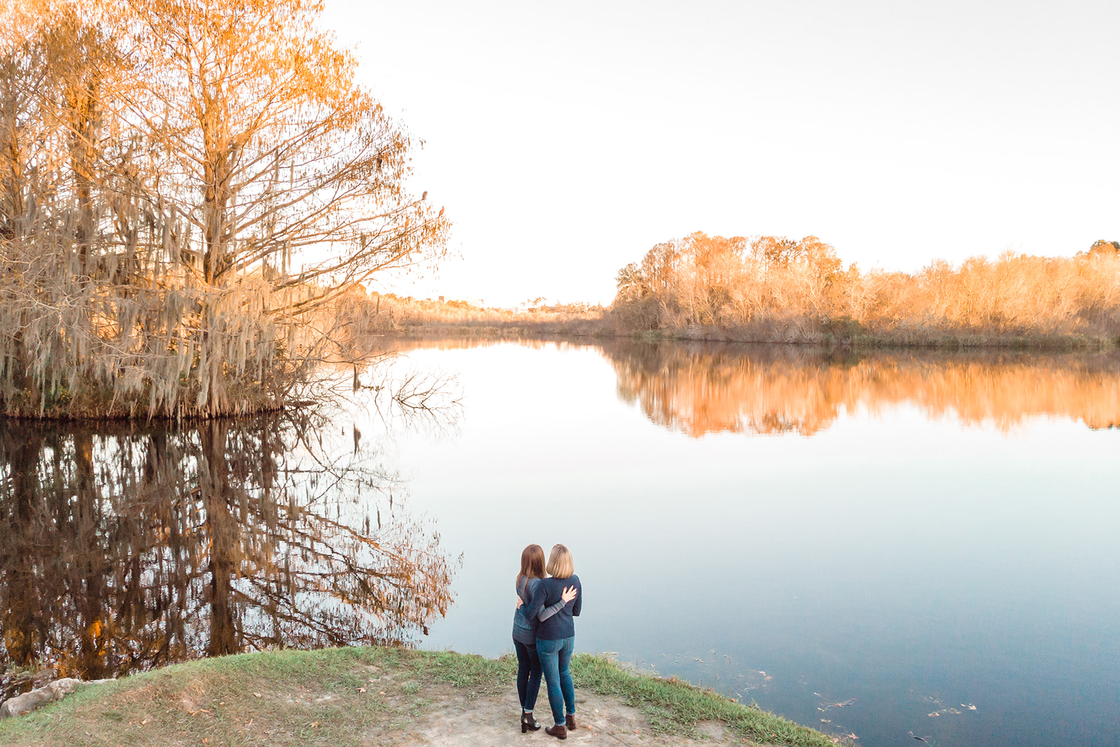 Two brides stand side by side, embracing by the edge of a serene lake, surrounded by the beauty of nature.
