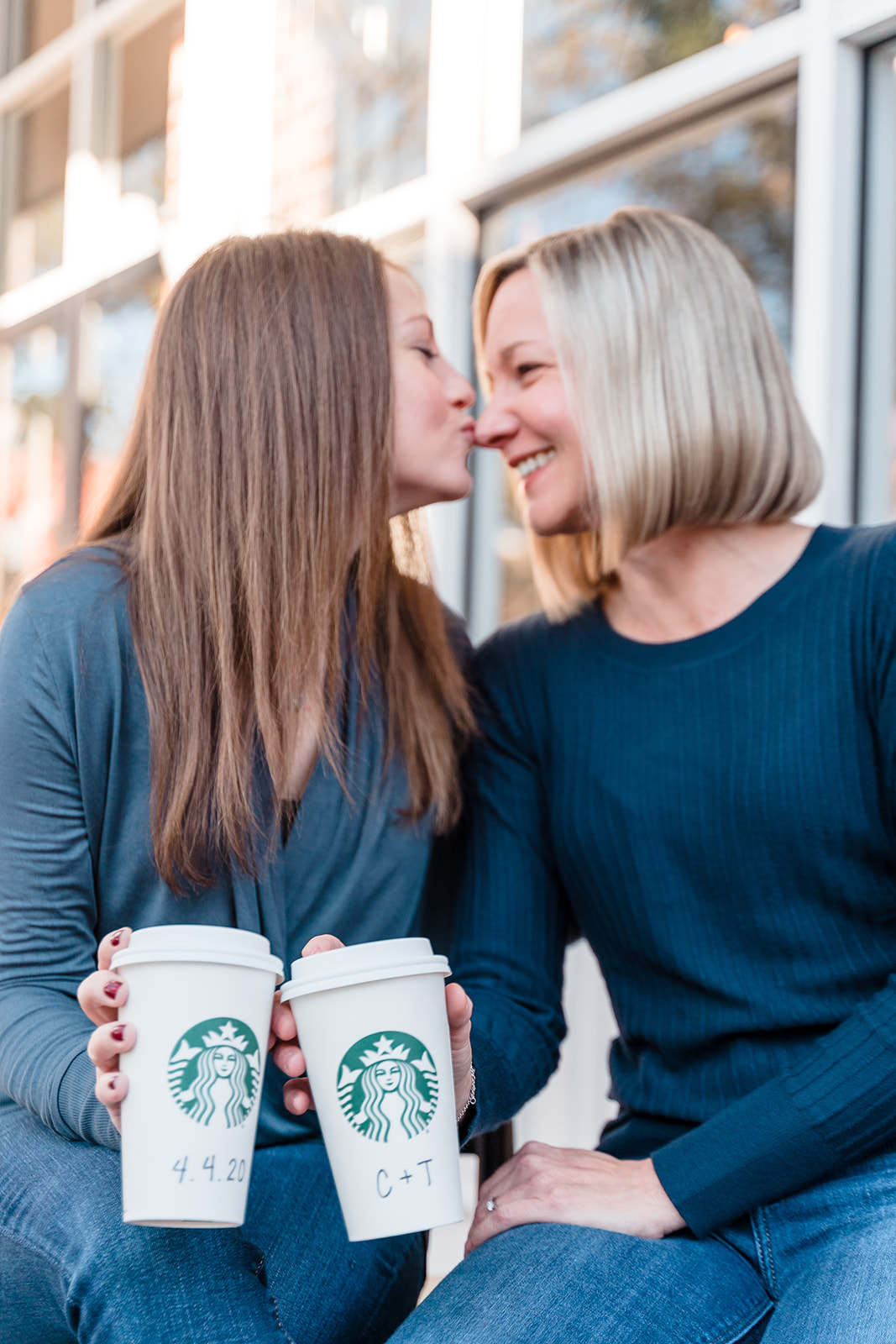 Two brides-to-be sit together, one kissing the other on the nose, while they hold Starbucks coffee cups adorned with their initials and the wedding date.
